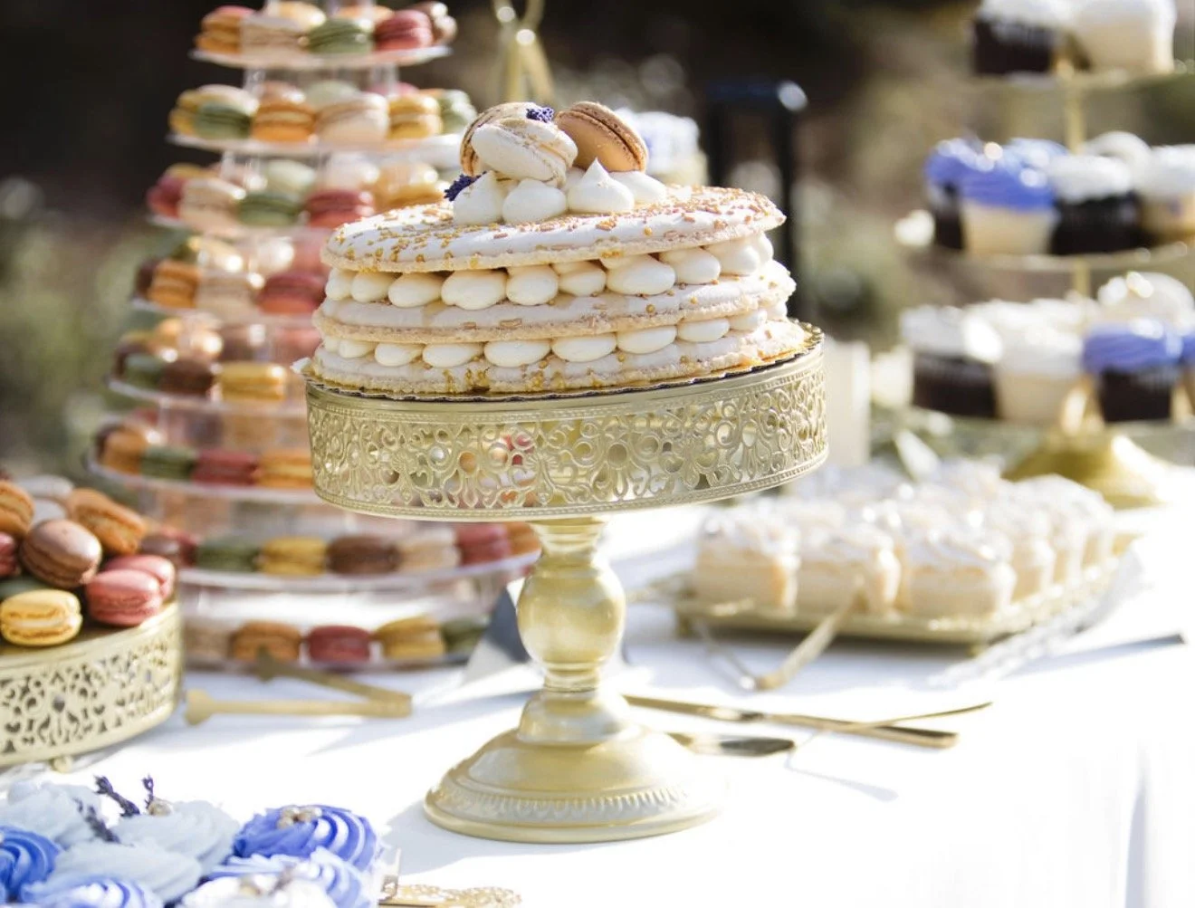 Elegant dessert table featuring a layered cake topped with macarons and cream, surrounded by colorful macarons and cupcakes.