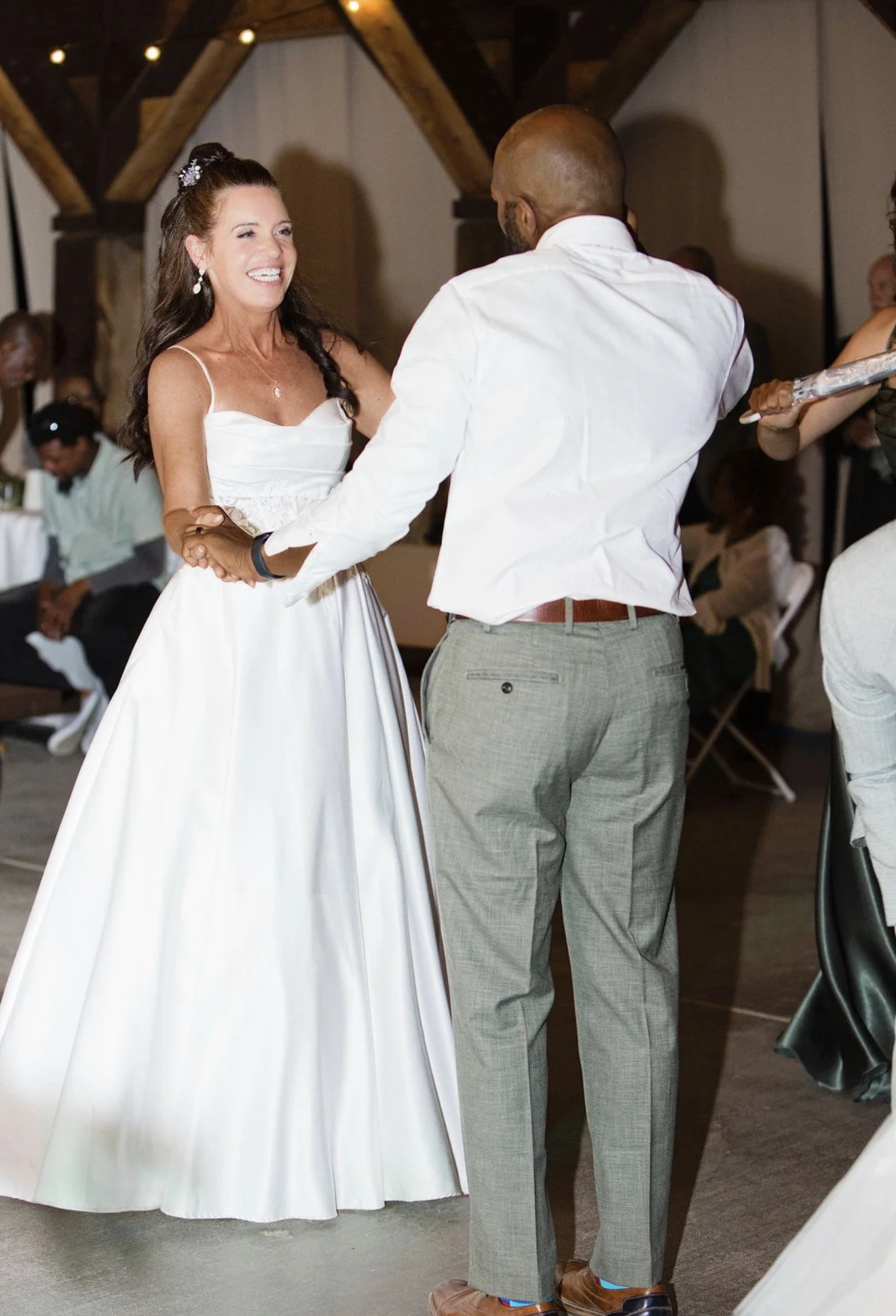 A bride and groom dancing at their wedding reception. The bride is smiling and wearing a white wedding dress, and the groom is dressed in a white shirt and gray pants.