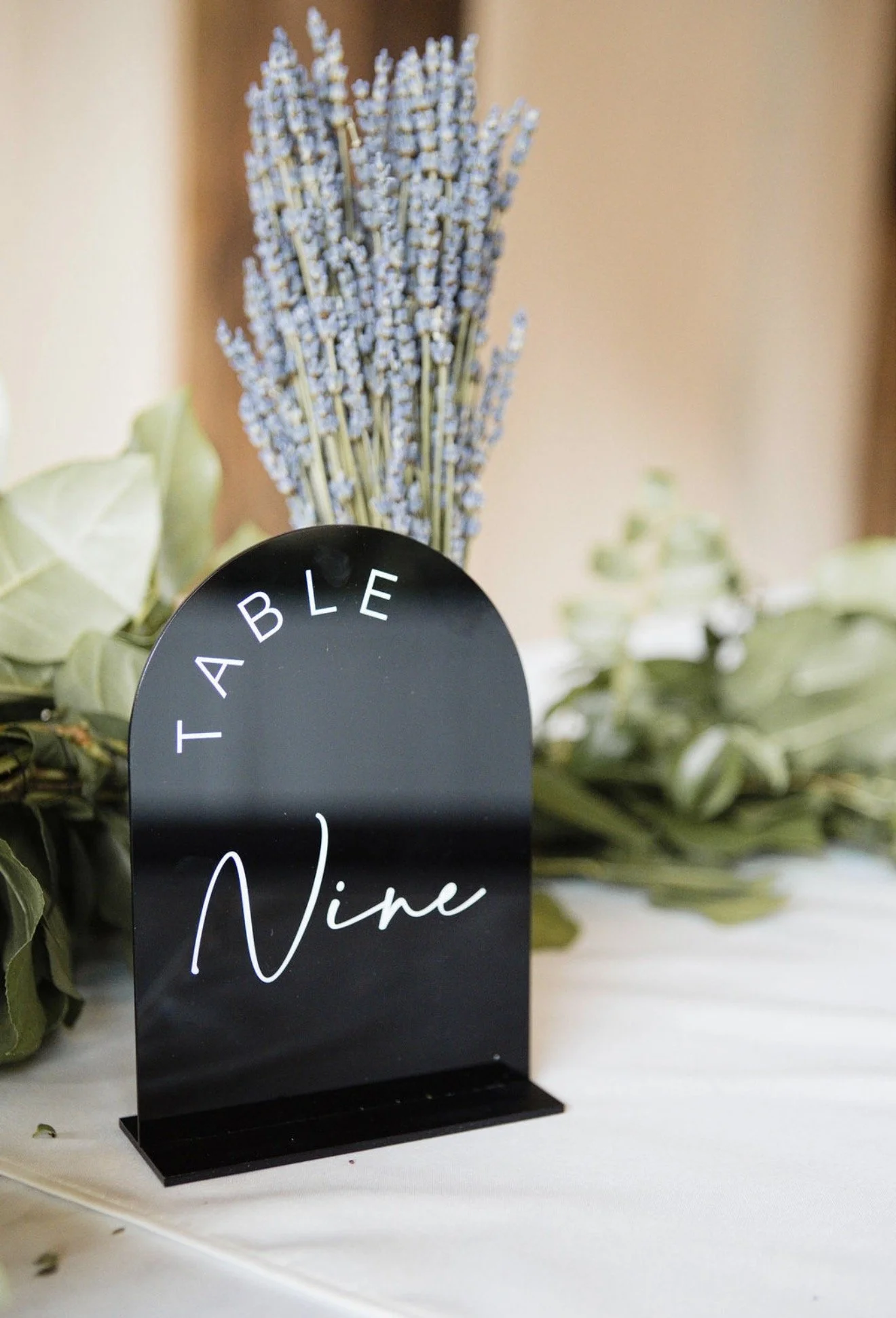 Black table number stand with white text displaying the number four, surrounded by green leaves and lavender flowers in the background.