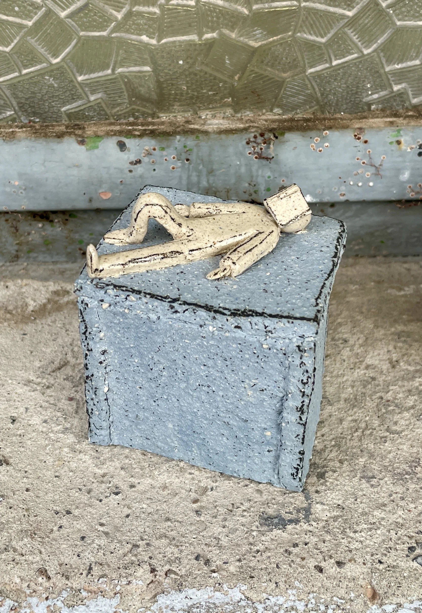 Handmade miniature ceramic sculpture of person laying down with an open book on its head on a square plinth.jpg