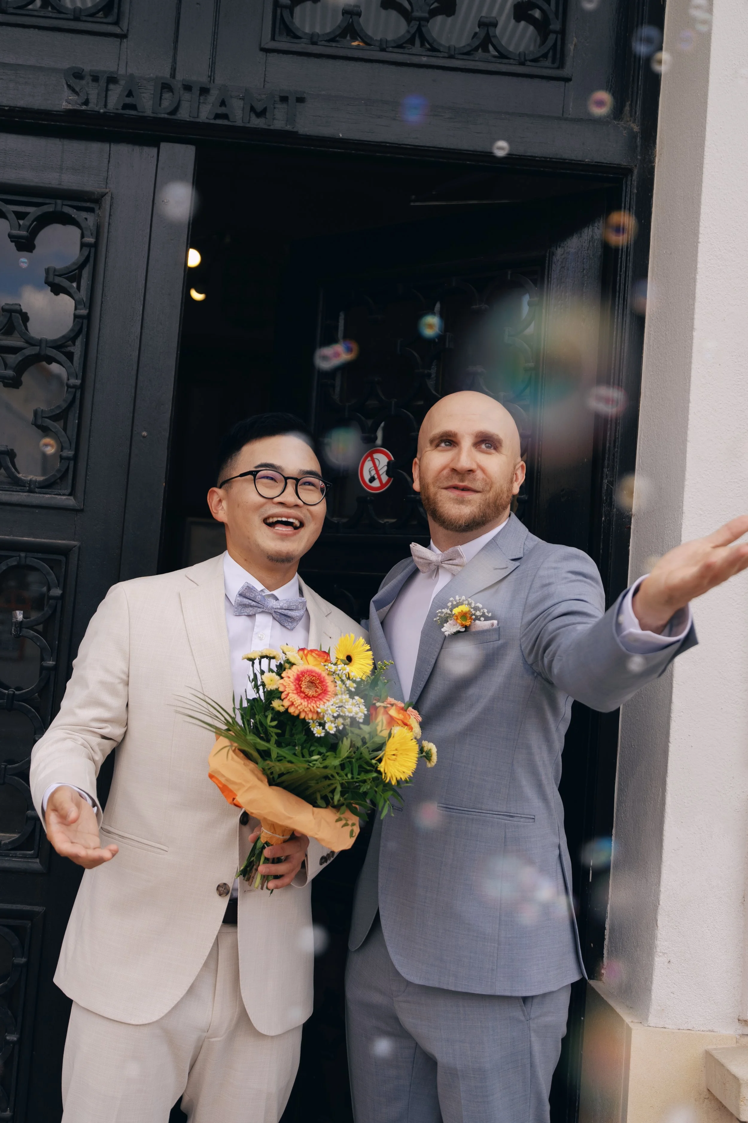 Two grooms celebrating outside the registry office with wedding bubbles.