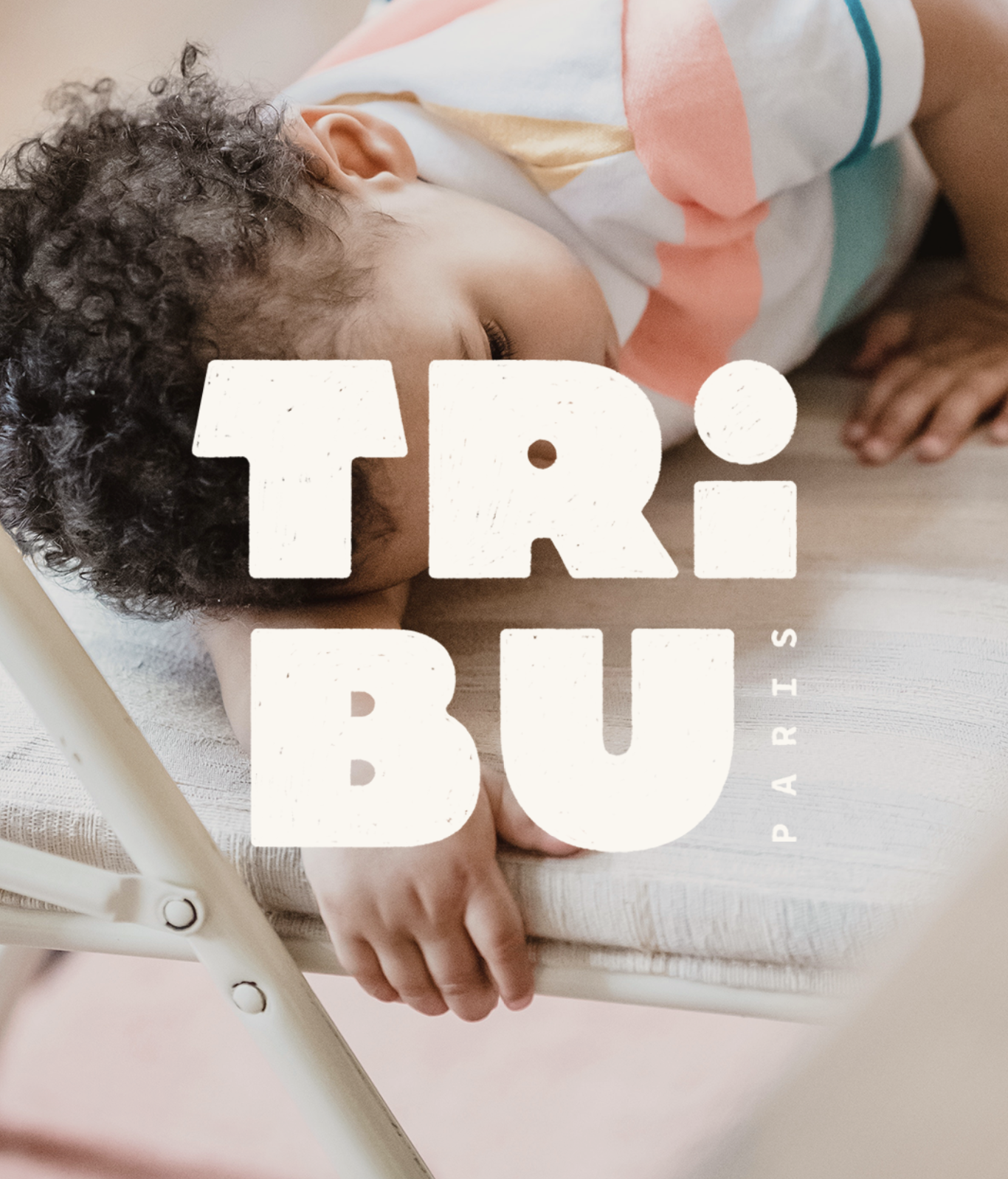 Un jeune enfant aux cheveux bouclés, allongé sur un lit, endormi, portant un t-shirt blanc avec des formes colorées. Sur l'image, en gros caractères blancs, il y a le texte "TRI.BU" avec le mot "PARIS" écrit en petit verticalement à droite.