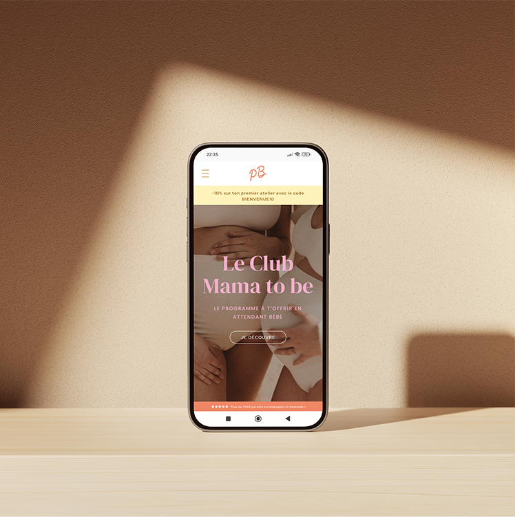 Téléphone affichant une page web pour un club de maternité, avec des images de femmes enceintes, sur une surface en bois contre un mur beige avec une lumière douce.