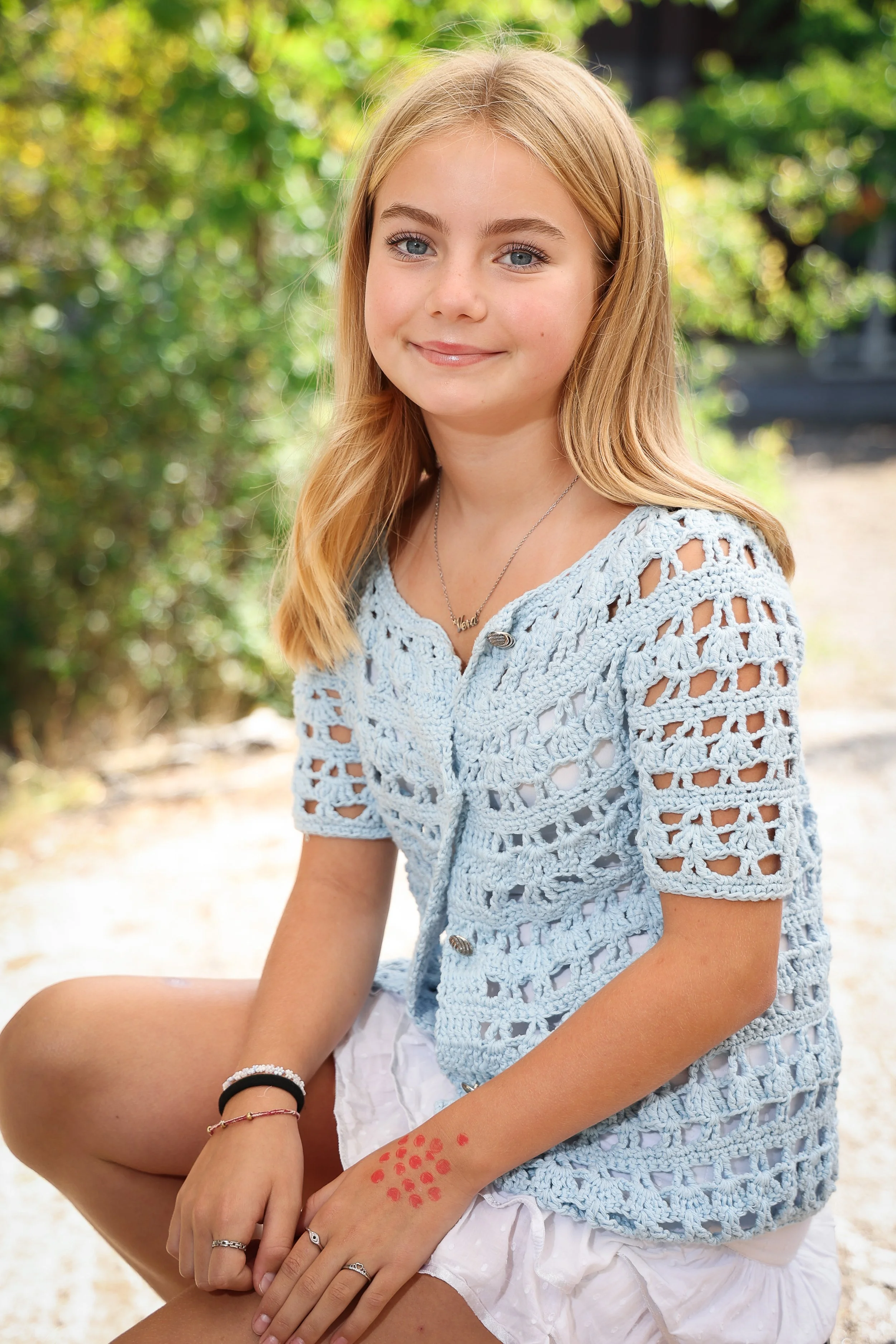 Ung flicka med blåa ögon, blond hår, som sitter utomhus i sommarväder, iklädd en ljusblå stickad kofta och vit kjol.