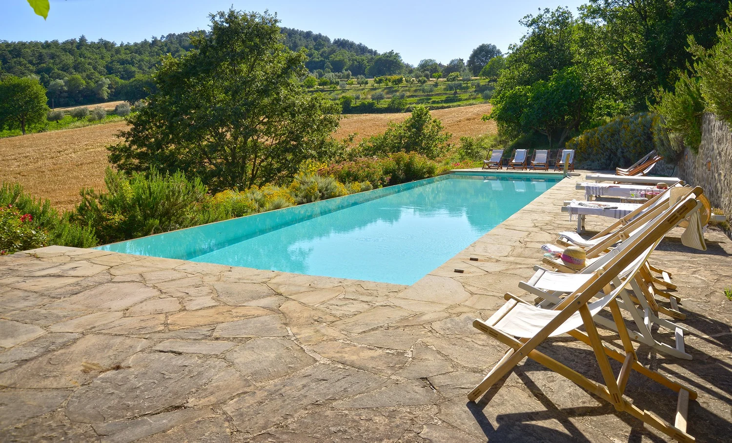 Infinity pool with a stone terrace and sun loungers overlooking fields and wooded hills.