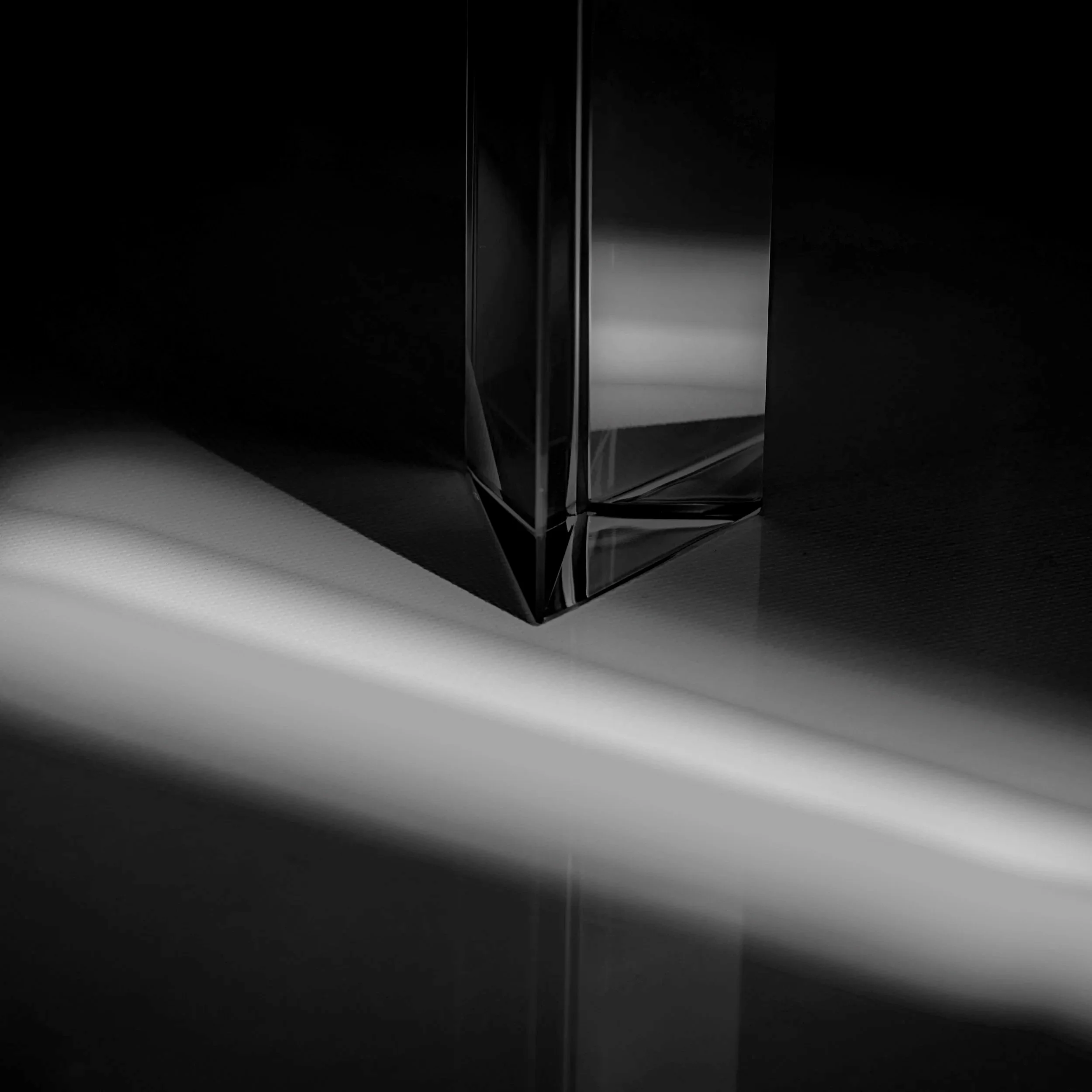 Close-up of a tall, transparent, glass or crystal rectangular object with sharp edges, on a reflective surface, with soft light creating reflections and shadows.