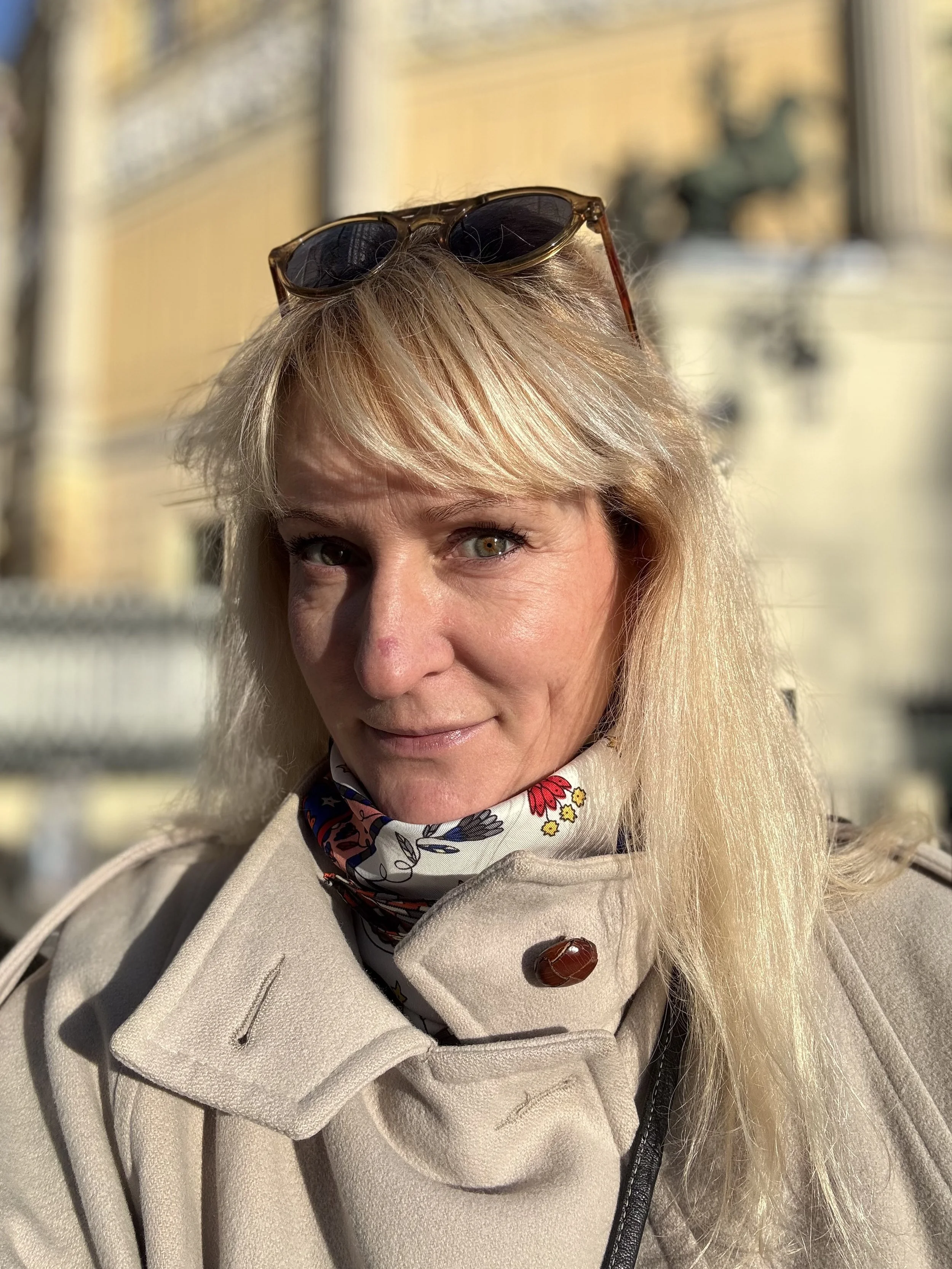 Close-up of a woman with blond hair, sunglasses on her head, wearing a beige trench coat and a floral scarf, outdoors in bright sunlight.