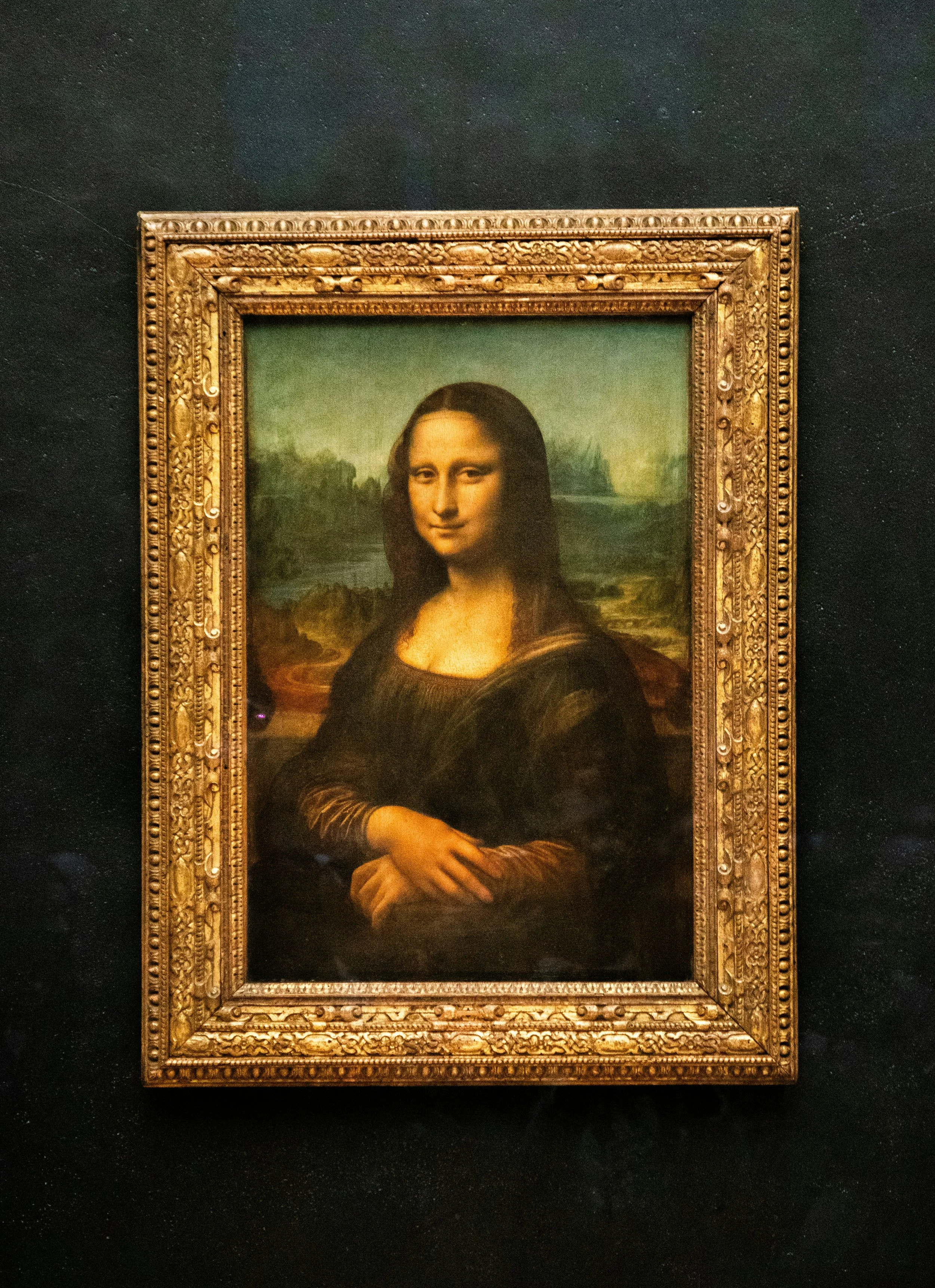 Famous portrait of Mona Lisa, a woman with dark hair, in front of a landscape with mountains and water, framed in an ornate gold frame.