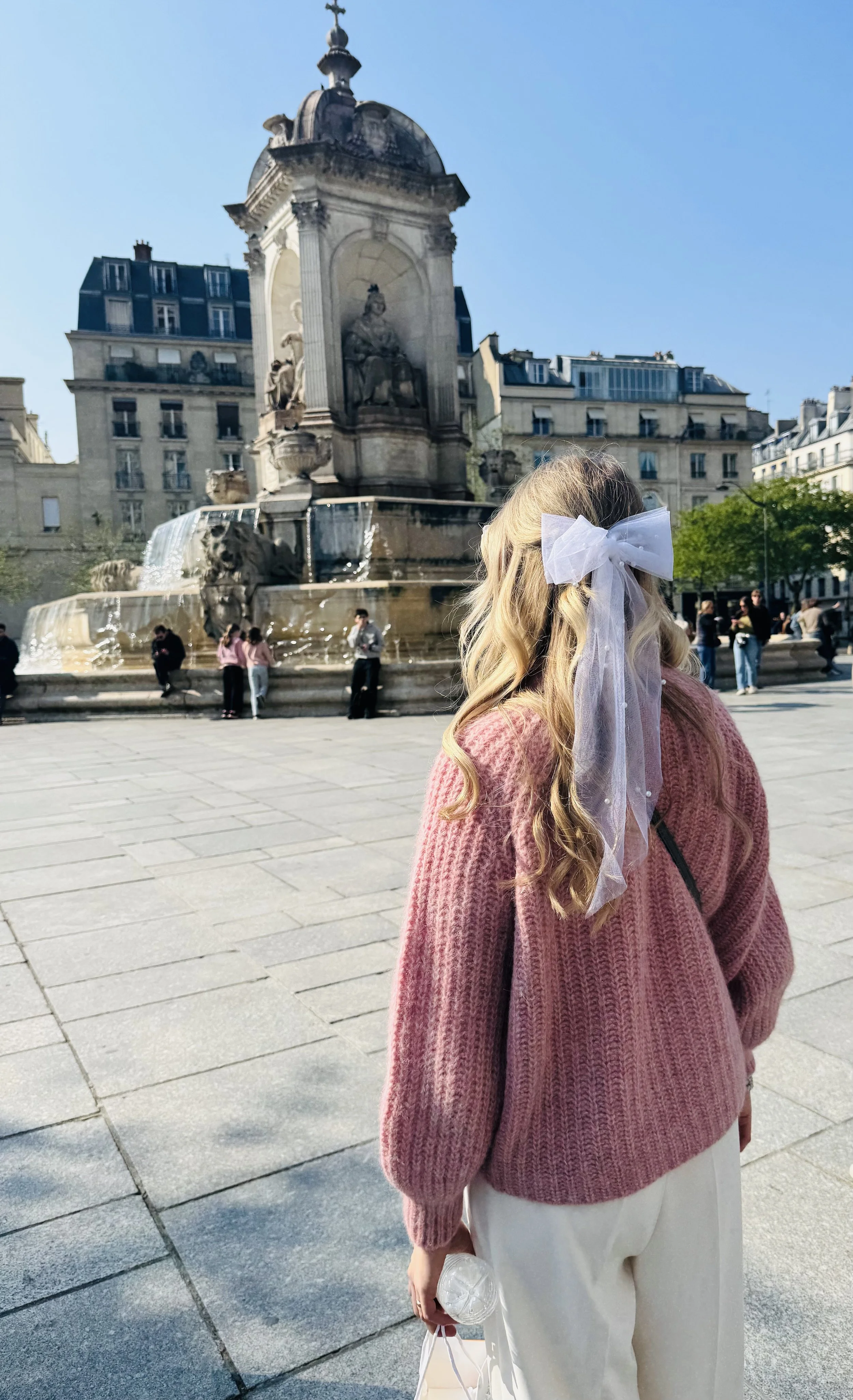 A woman with long blonde hair, wearing a pink sweater and white pants, stands in a city square with a fountain featuring sculptures and water in the background. She holds a small clear bag and has a large white bow in her hair.