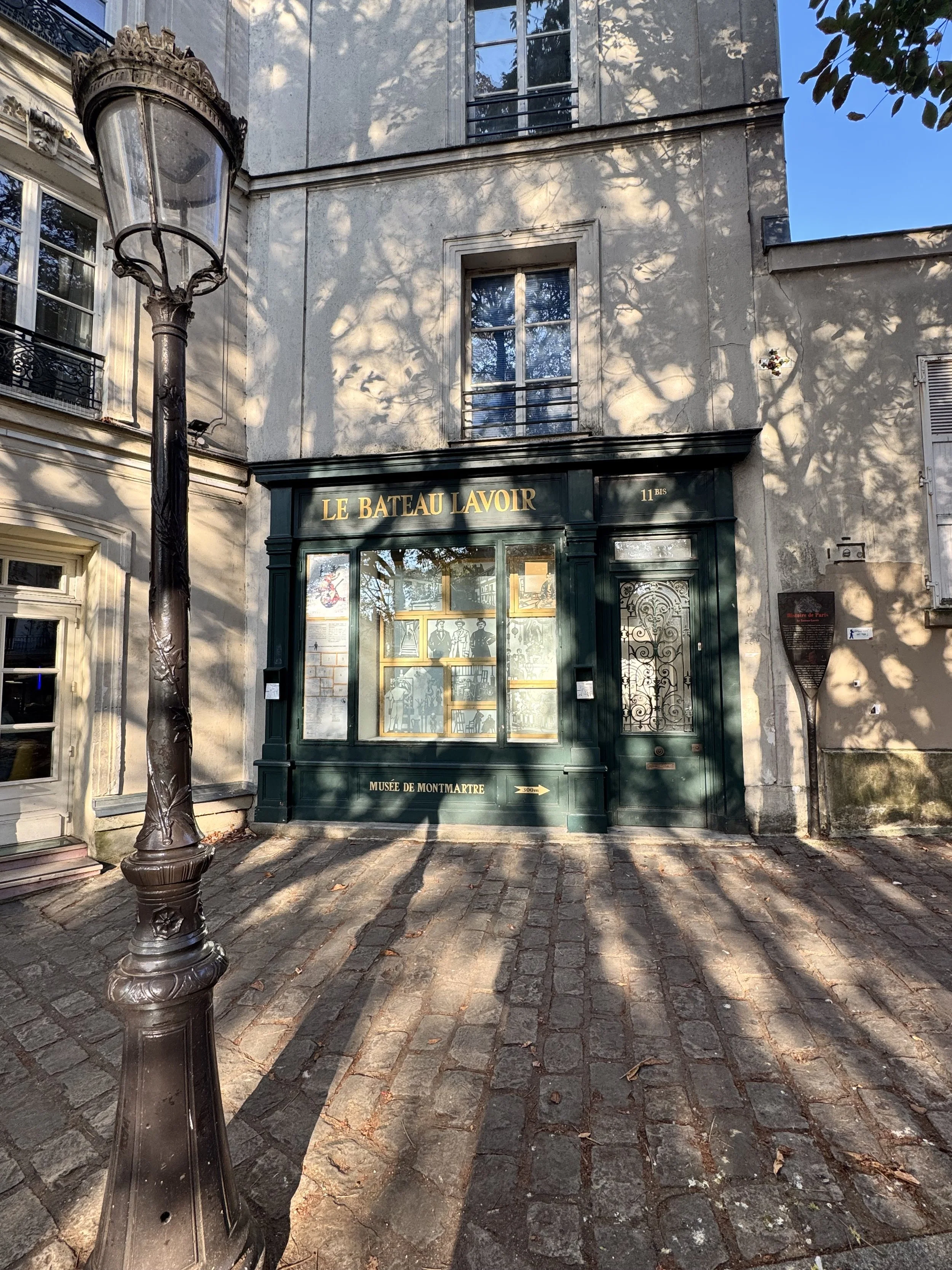 A street view showing a building with a dark green storefront labeled 'Le Bateau Lavoir' and 'Musée de Montmartre,' with a lamp post in the foreground and trees casting shadows on the building. The street is cobblestone.