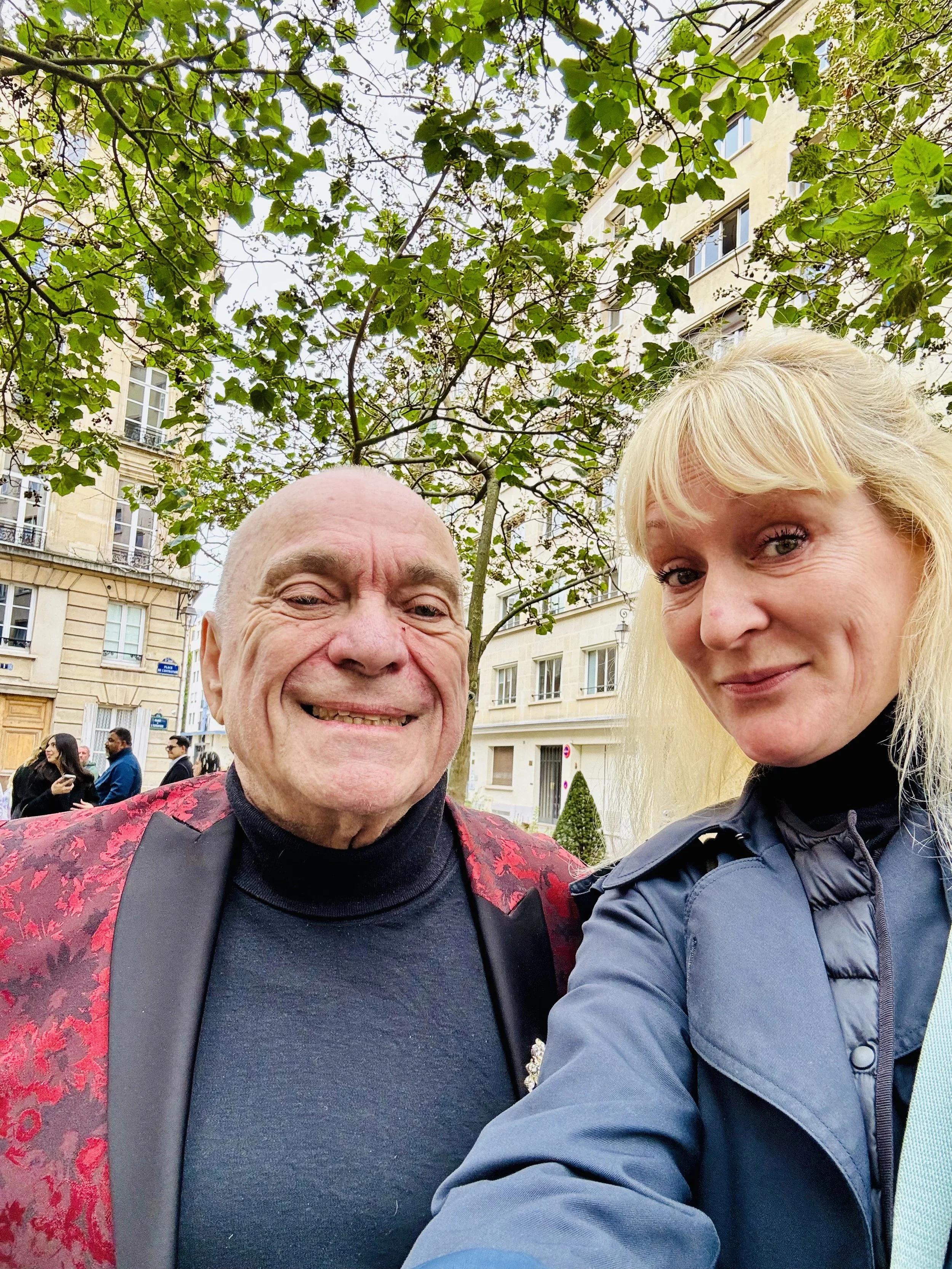 A selfie of a man and a woman outdoors in an urban setting with trees and buildings in the background.