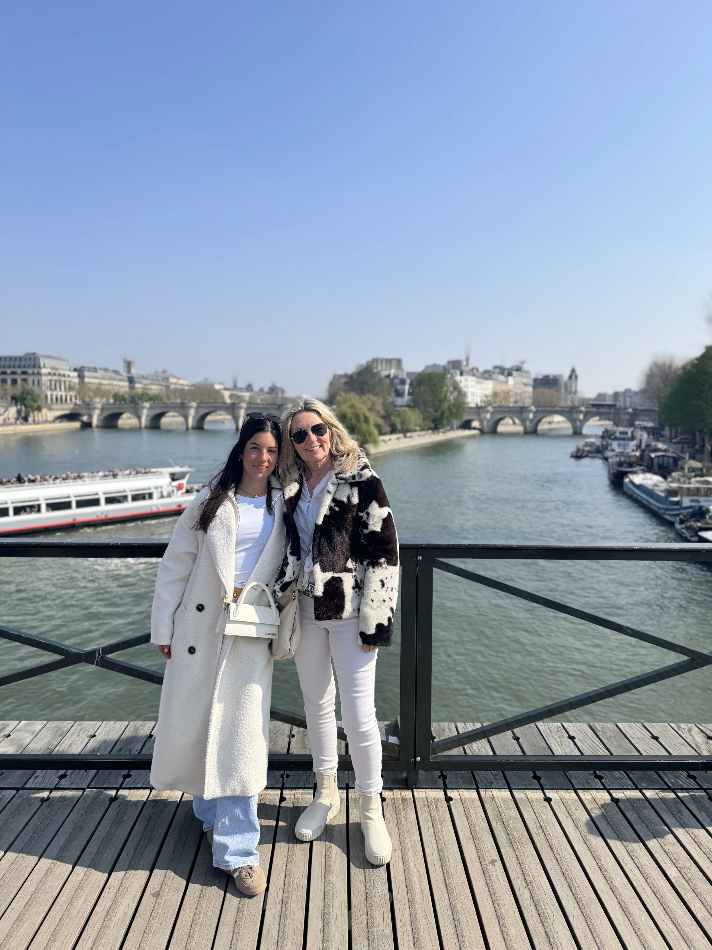 Two women standing on a wooden bridge with the Seine River and Parisian buildings in the background. One woman has long dark hair and is wearing a long white coat, blue jeans, and beige shoes. The other has blonde hair, sunglasses, a black and white 