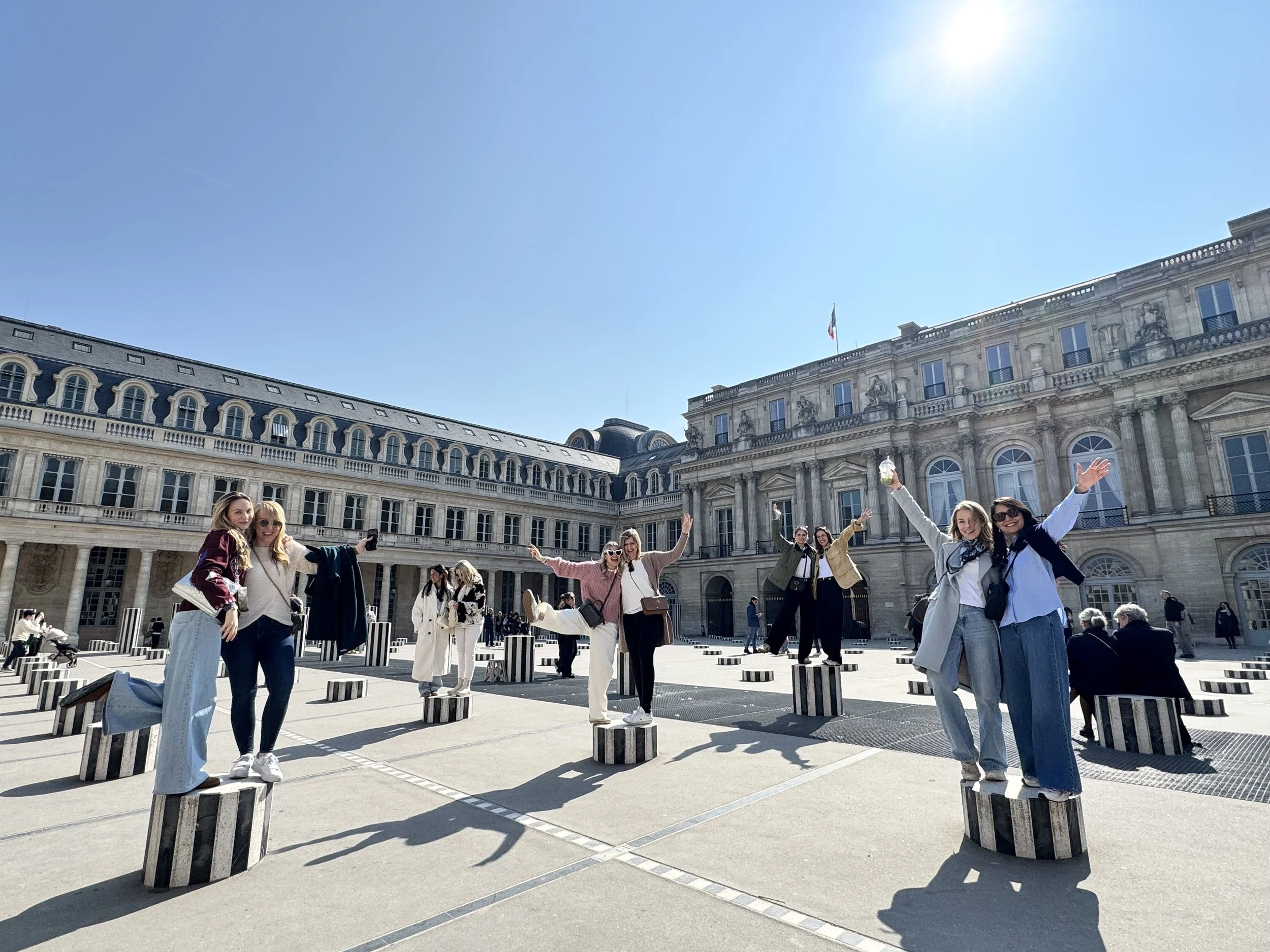Group of women posing on striped cylindrical objects in a city square with historic buildings in the background, under a clear blue sky.