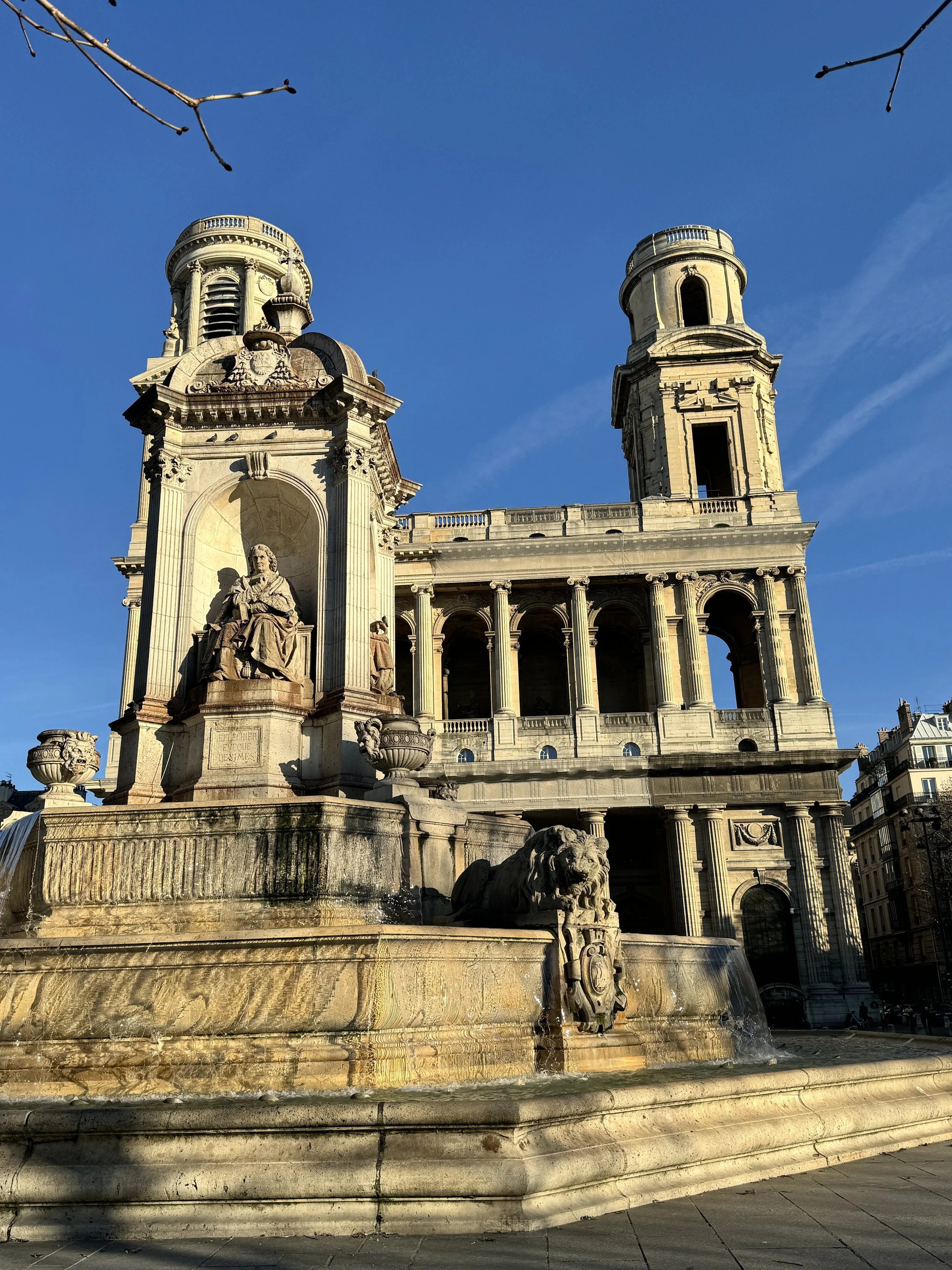 A historic stone fountain with sculptures and water feature, in front of a large building with two towers and columns, under a clear blue sky.