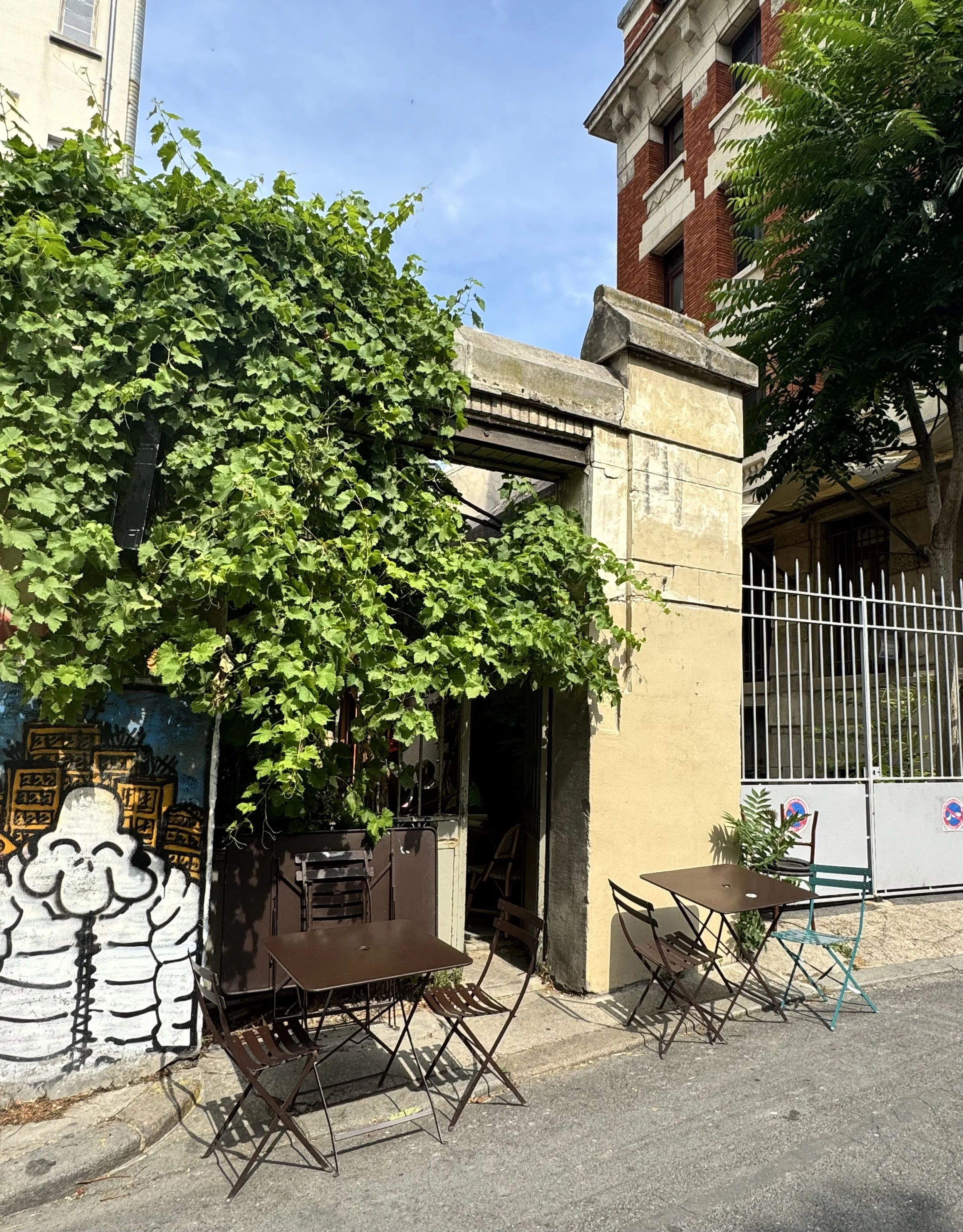 Außenbereich eines Cafés mit mehreren Tischen und Stühlen, grün belaubte Bäume, eine Mauer mit Graffiti-Kunst, ein Tor sowie eine mehrstöckige rote Ziegelbau
