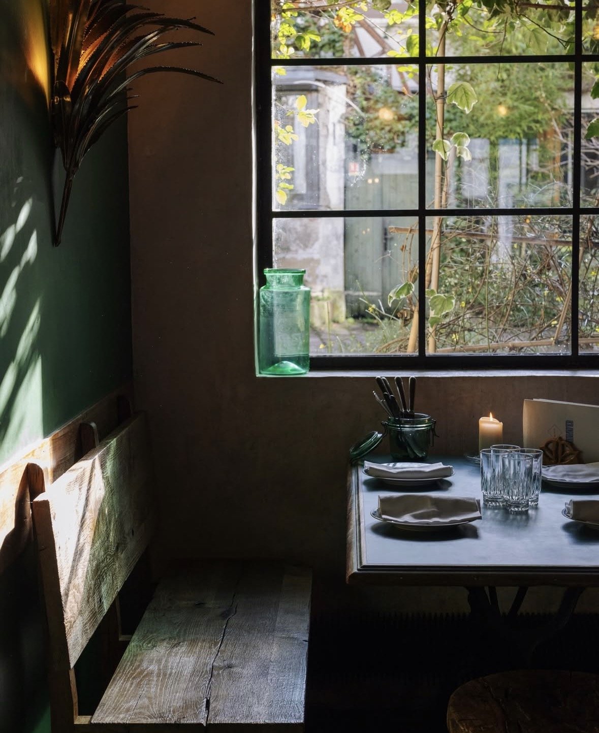 Ein Esstisch mit abgestellten Tellern, Gläsern, Besteck, einem Kerzenlicht und einem schwarzen Ständer. Im Hintergrund ein Fenster mit Blick auf einen Garten mit Pflanzen und einer mehrfarbigen Mauer.
