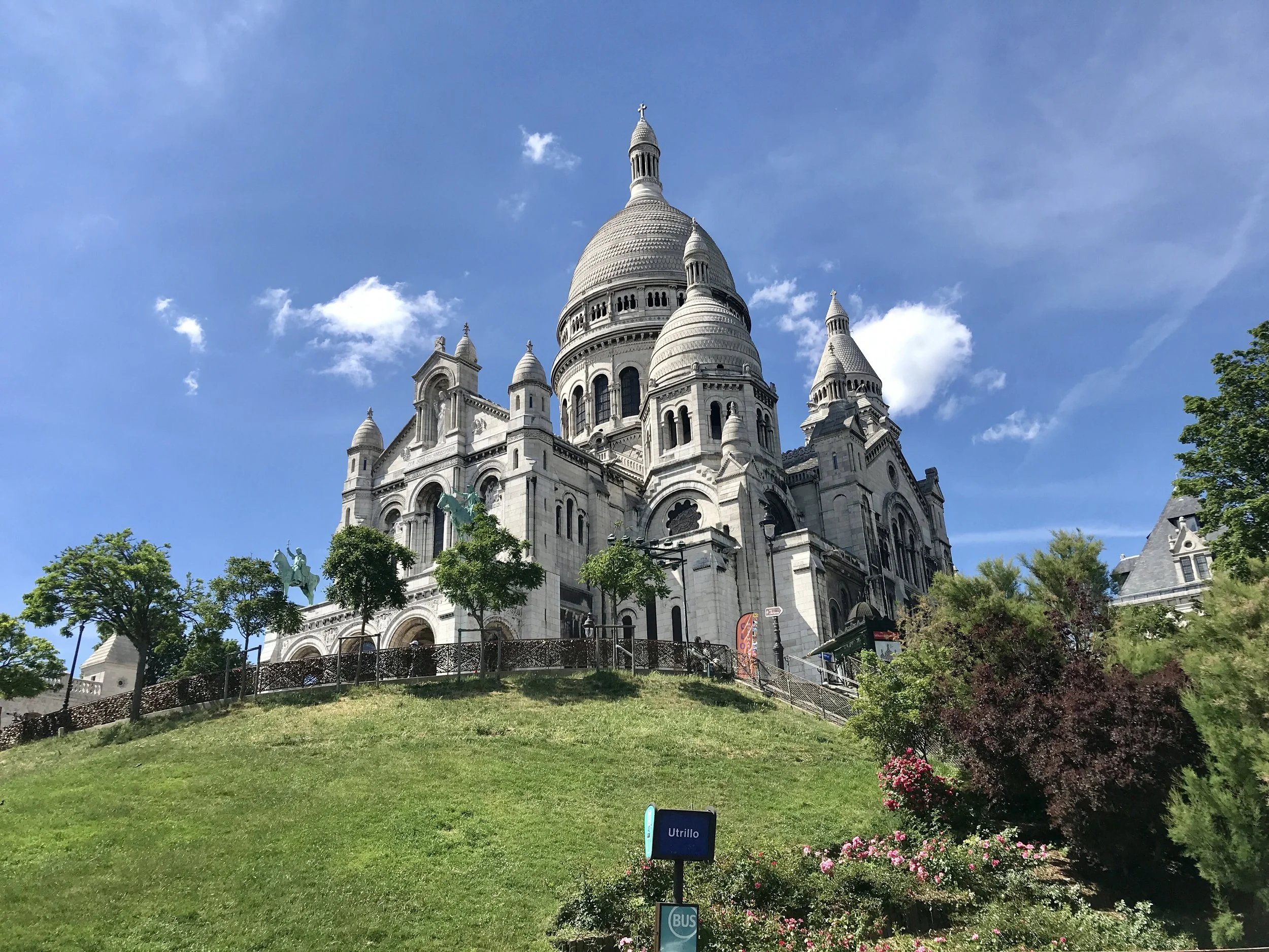The Sacré-Cœur Basilica, a white stone church with multiple domes and towers, situated on a hill with green grass and trees in the foreground, under a partly cloudy blue sky.