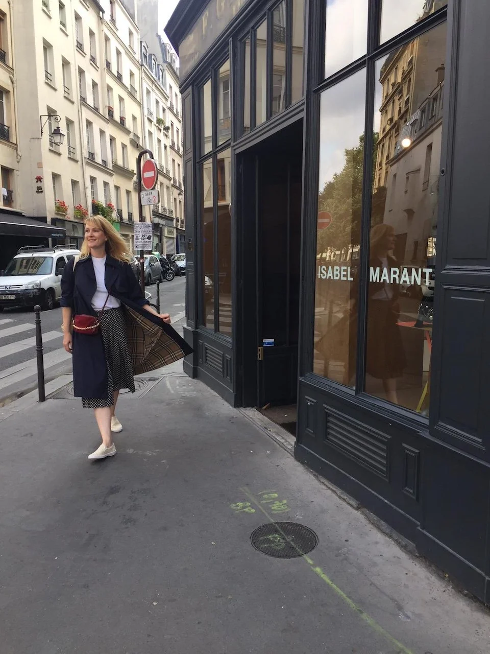 A woman walking on a city sidewalk in front of a black storefront with the name 'Isabel Marant' on the window. She is smiling and wearing a navy coat over a white top, patterned skirt, and white sneakers, carrying a red purse with a checkered lining. Parked cars, buildings, and street signs are visible in the background.