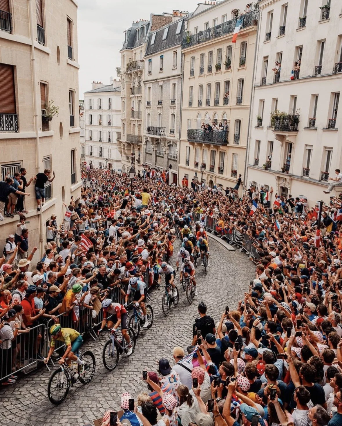 A crowded street in Paris with spectators watching a cycling race through the narrow, cobblestone street, with apartment buildings on both sides and French flags hanging from balconies.