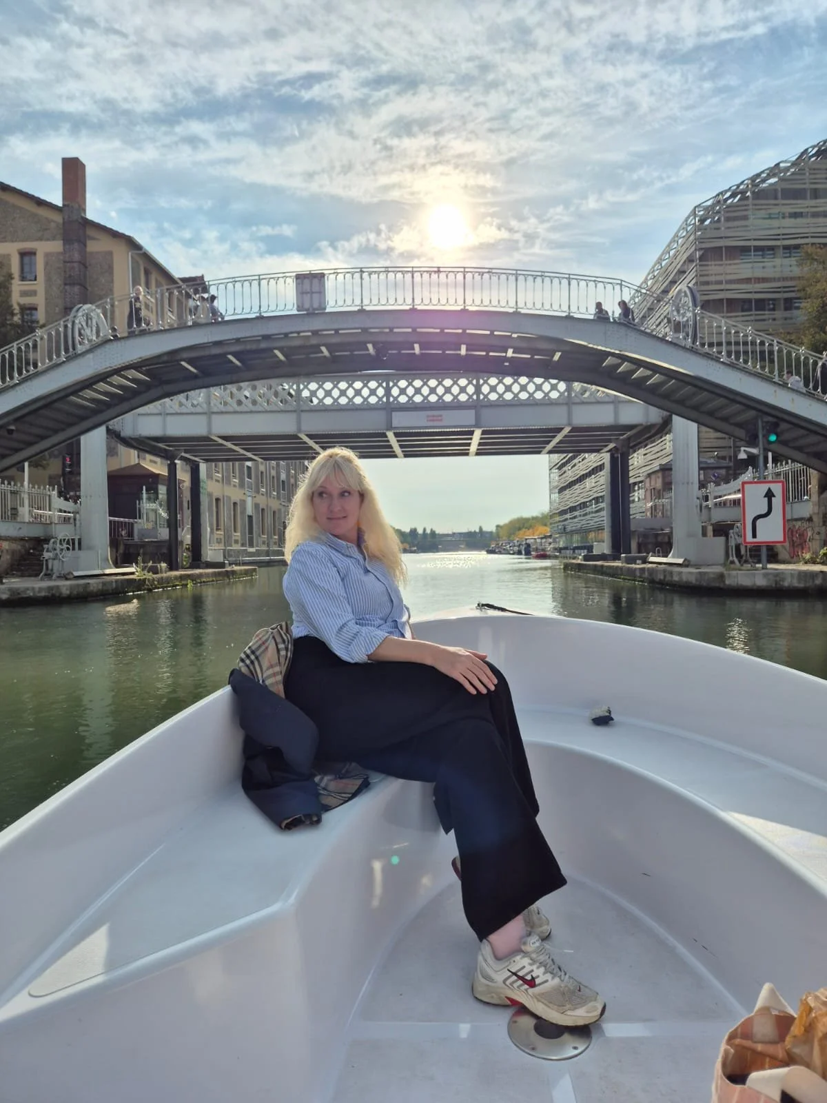 A woman sitting on a boat on a canal, with a white arched bridge overhead and buildings on either side, under a sky with scattered clouds and the sun shining.