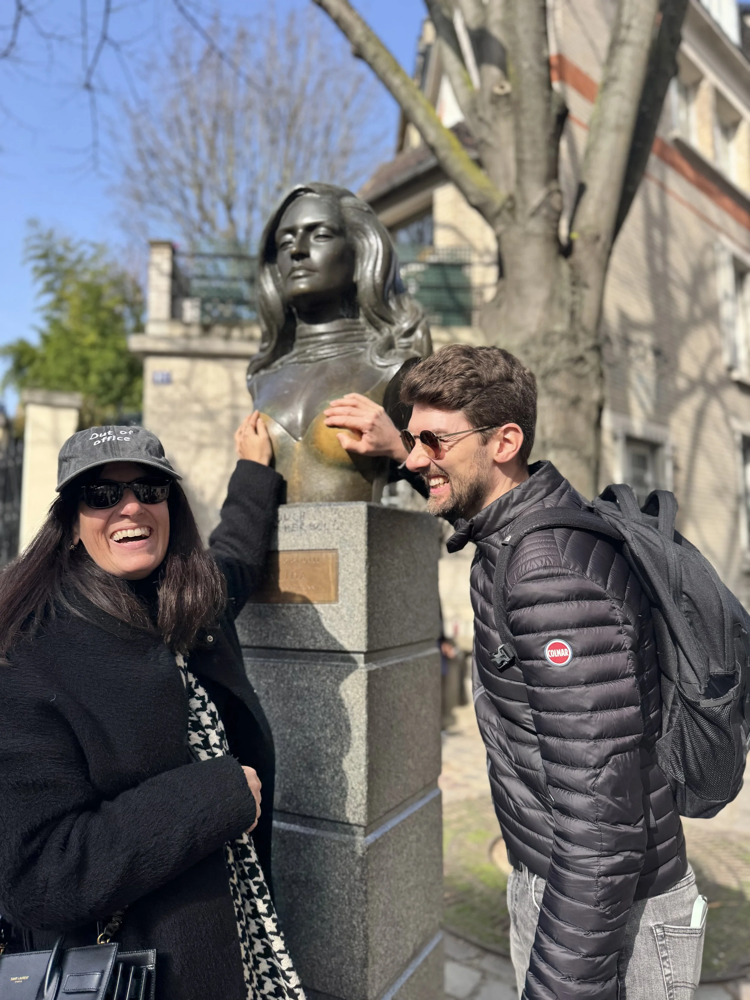 Two people, a woman and a man, smiling and posing by a bust sculpture of a woman in an outdoor setting with trees and buildings in the background. The woman is wearing sunglasses, a hat, and a black coat, while the man is wearing sunglasses, a black 