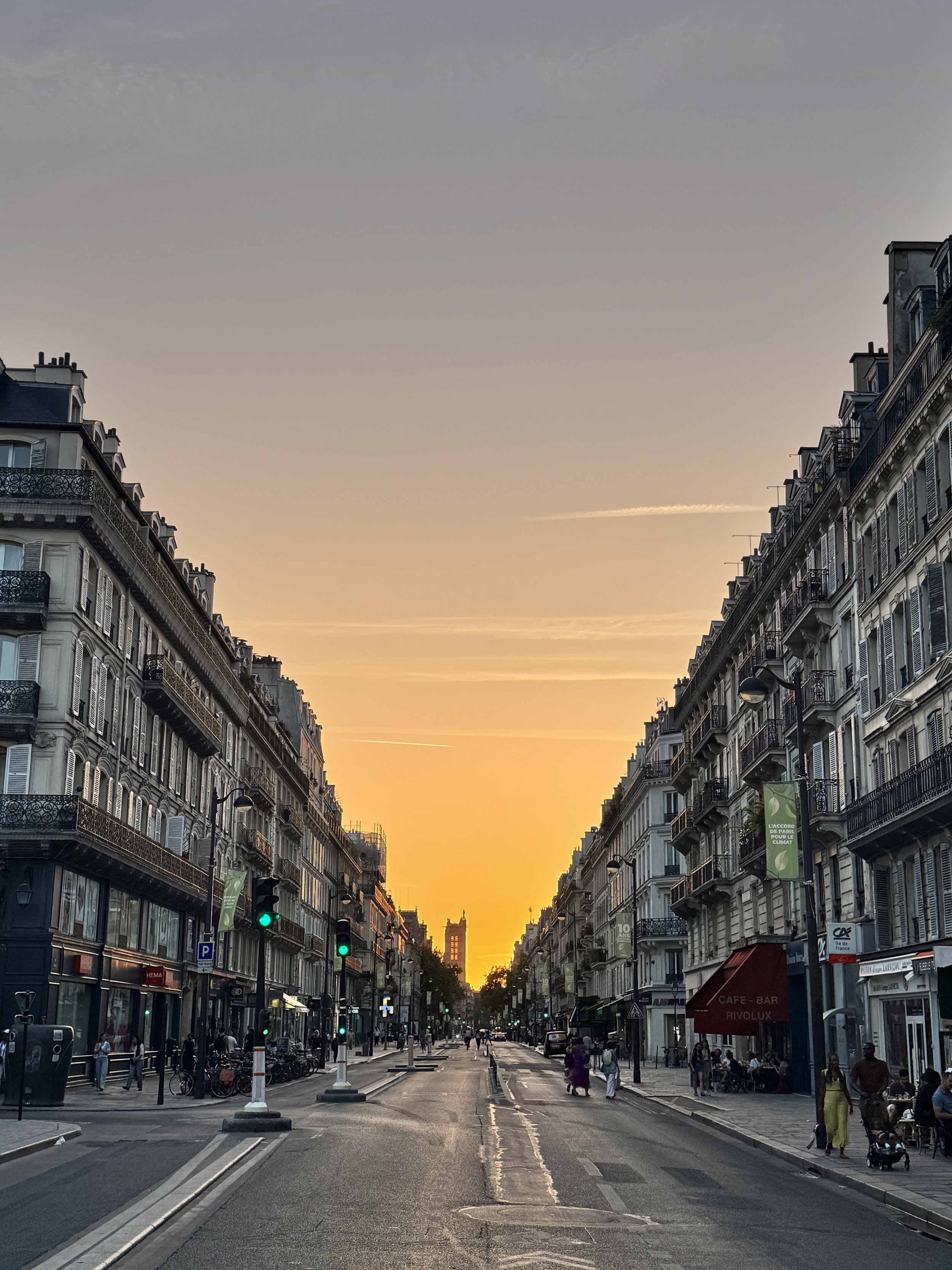 Straßenansicht in Paris während des Sonnenuntergangs mit historischen Gebäuden, Fußgängern, Straßenverkehr und einem Himmel in sanften orange- und grautönen.