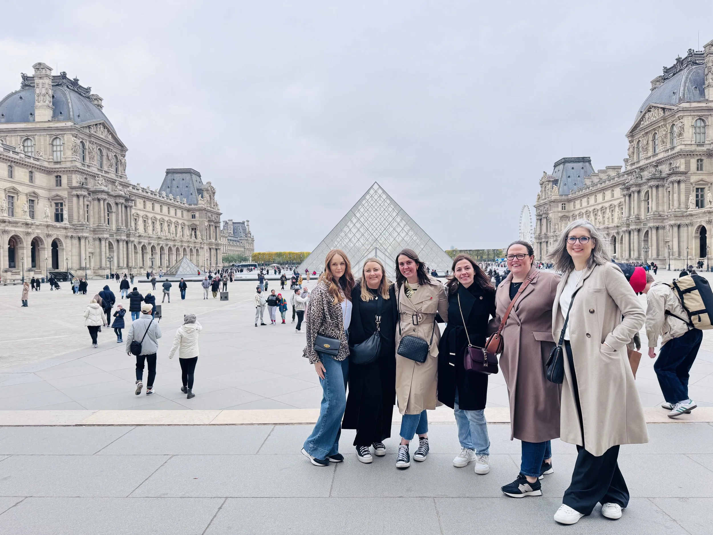 Six women standing together in front of the Louvre Museum with the glass pyramid in Paris, France.