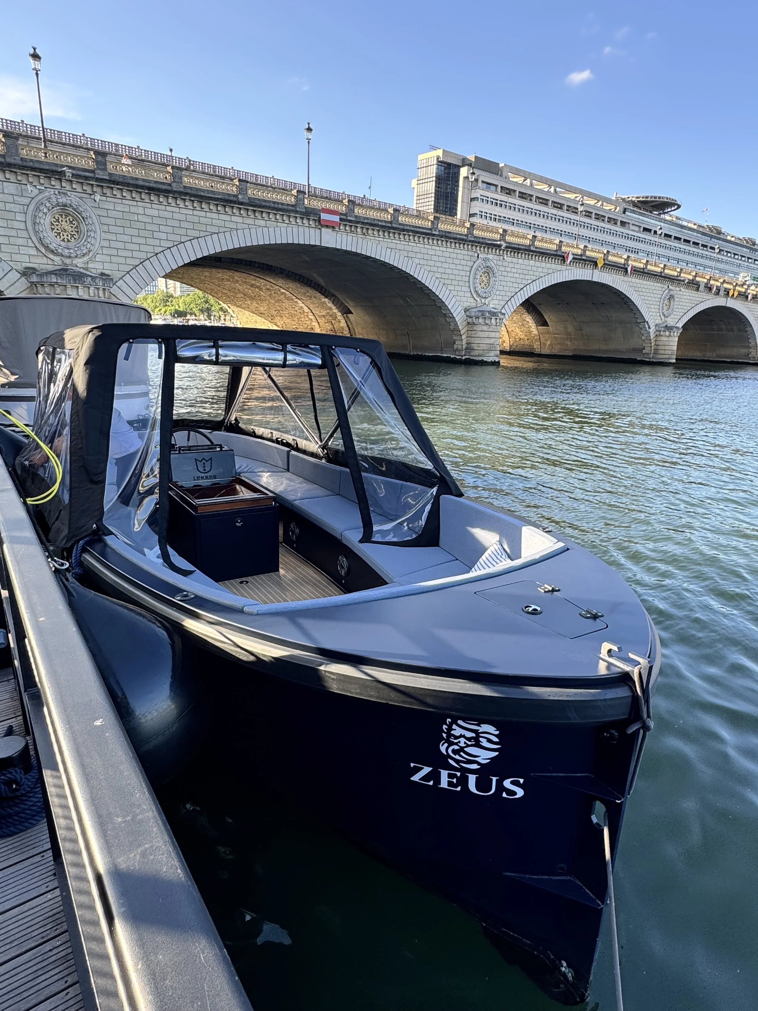 Ein Boot mit dem Logo 'ZEUS' liegt an einer Anlegestelle auf einem Fluss oder See. Im Hintergrund ist eine Brücke mit mehreren Bögen und Gebäuden zu sehen, die bei Sonnenlicht erstrahlen.