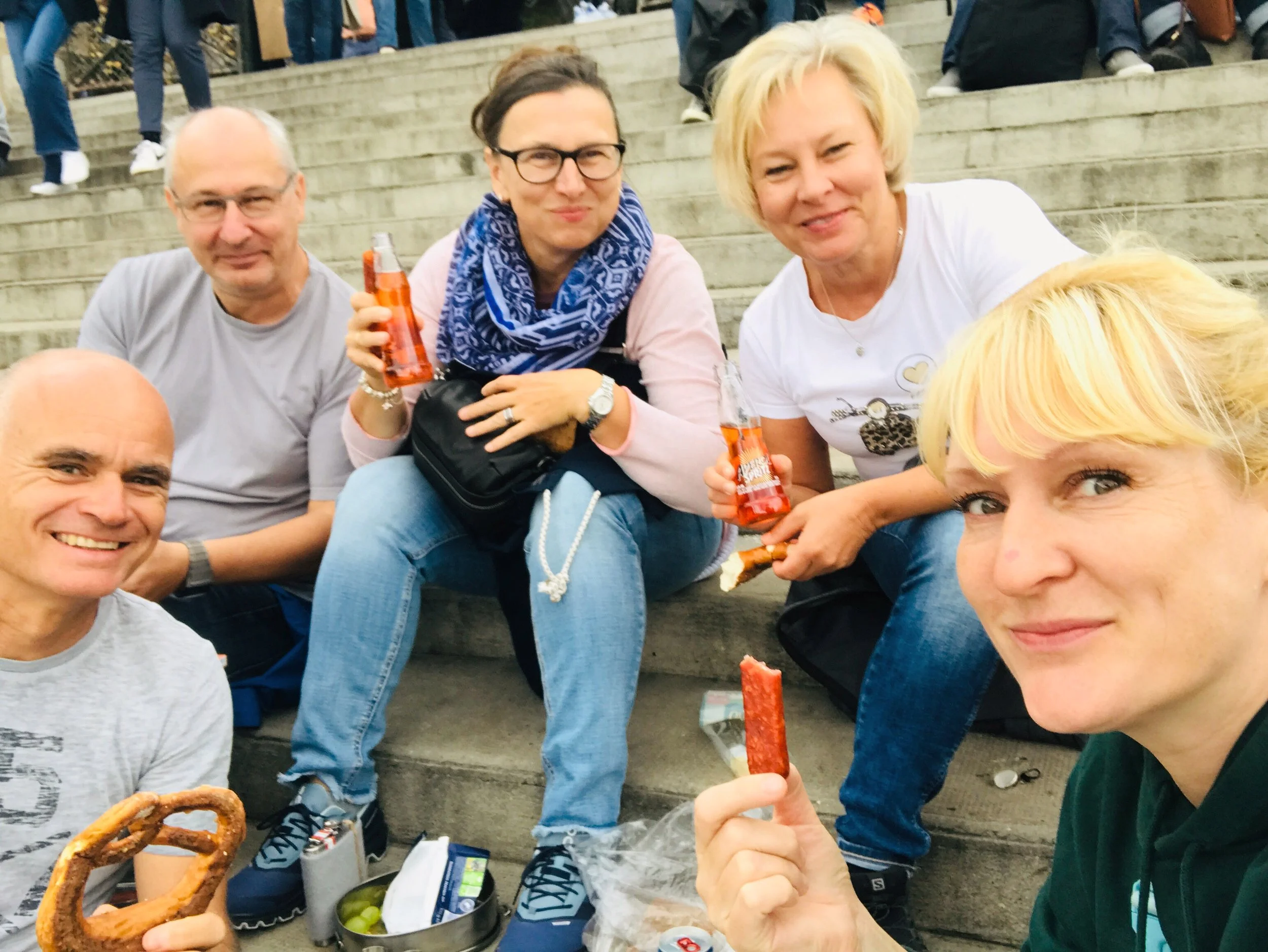 Group of five people sitting on outdoor steps, holding bottles of orange drinks and snacks, smiling at the camera during a casual gathering.
