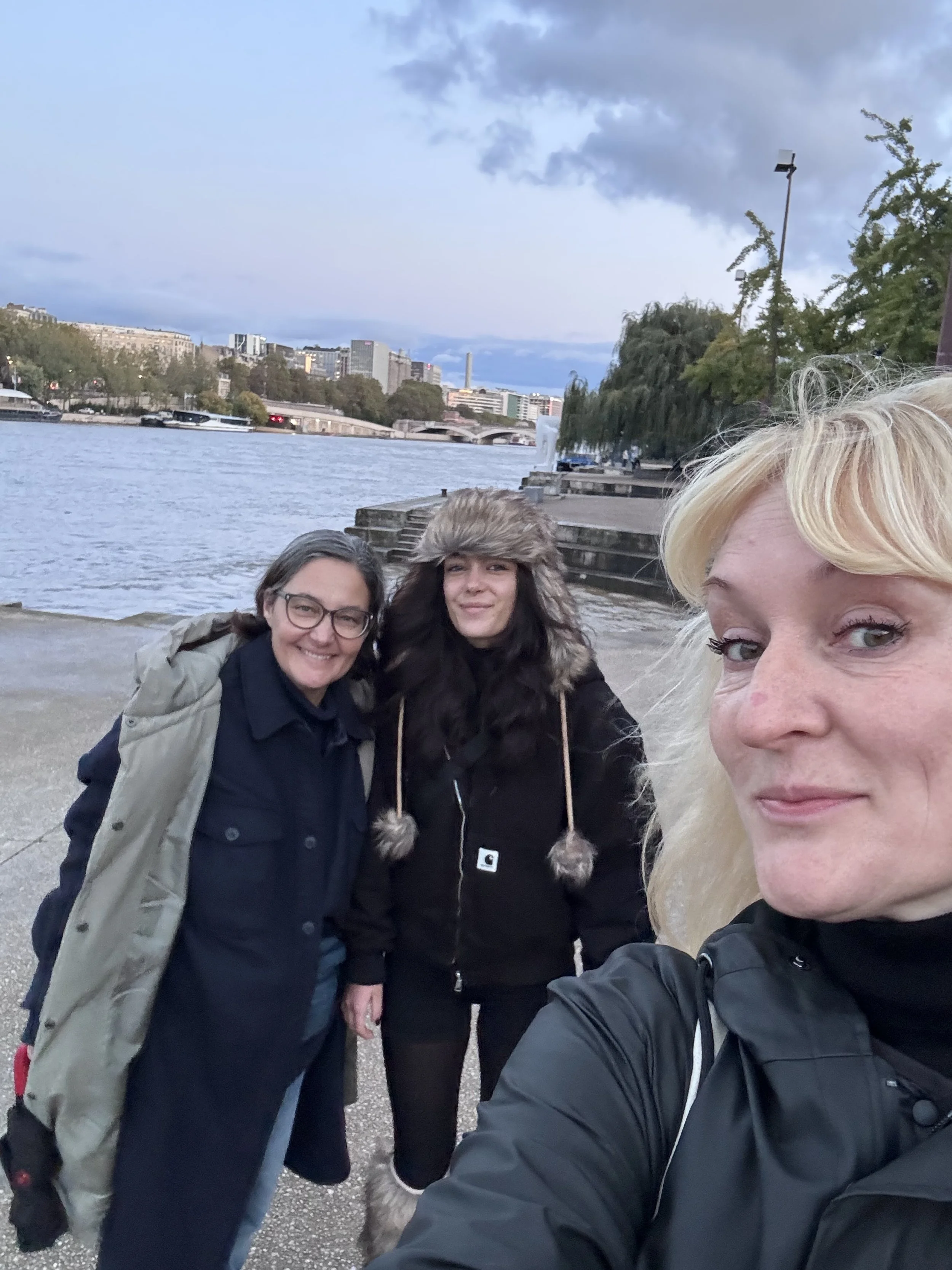 Three women taking a selfie by a river in an urban area, with city buildings and trees in the background during dusk.