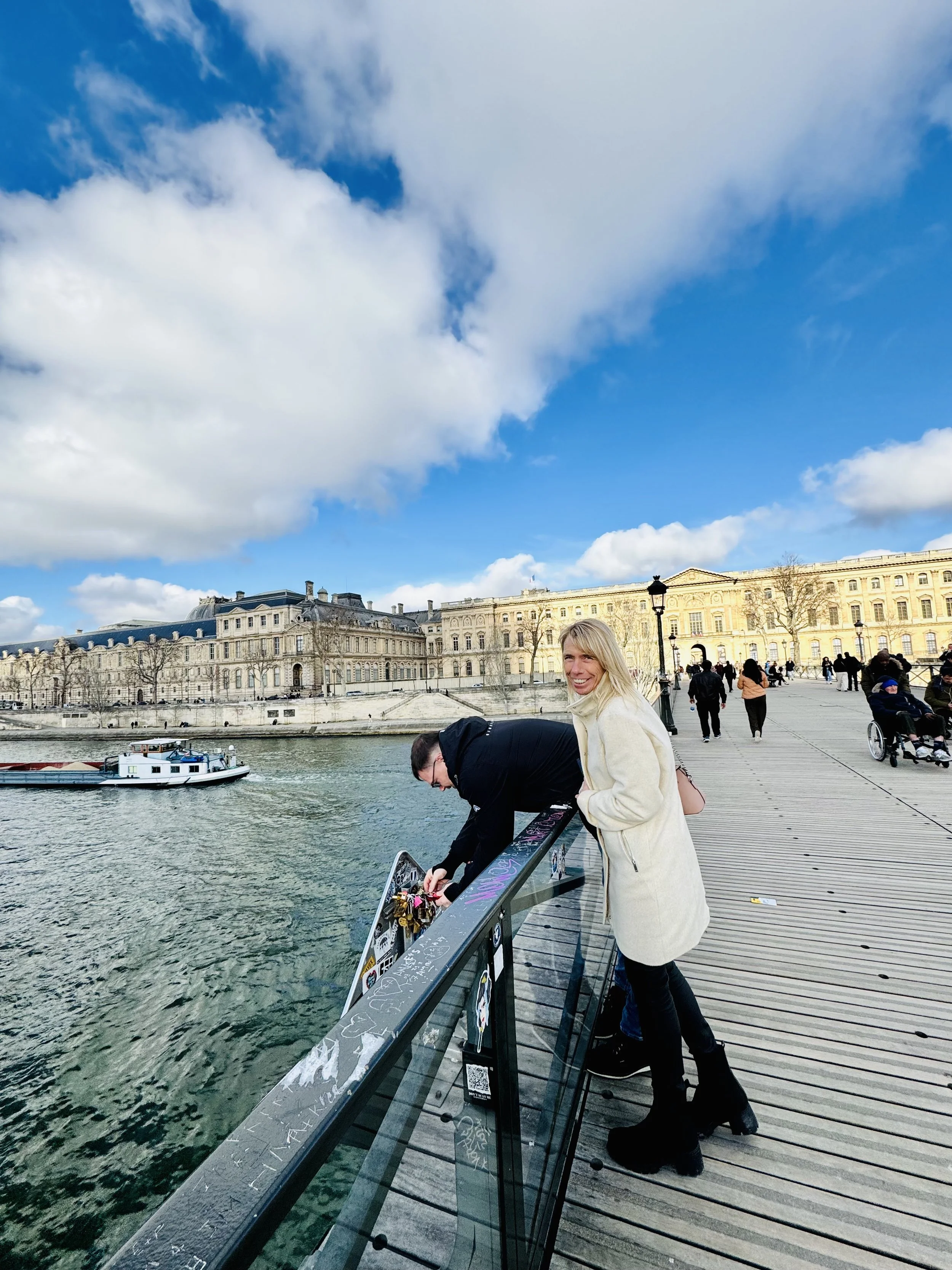 Two people on a wooden bridge over a river in Paris, France, with historic buildings in the background. One woman is smiling at the camera while the other is adjusting or placing locks on the bridge's railing. The sky is partly cloudy with blue skies