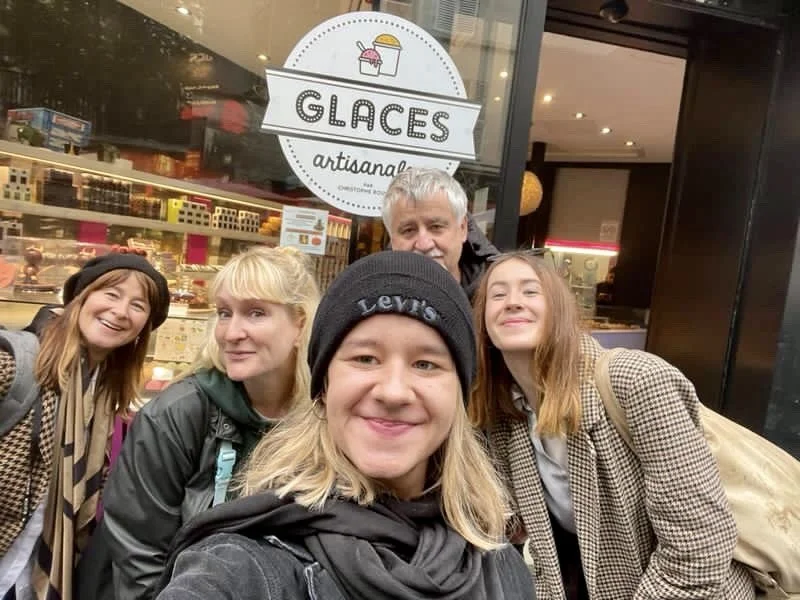 Group of five people taking a selfie outside a gelato shop with a sign that says 'Glaces artisanaux' in French, indicating an ice cream shop.