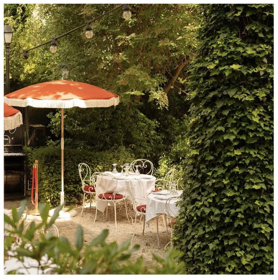 Ein Garten mit gedecktem Essbereich, weißen Tischen mit weißen Tischdecken, roten Stühlen mit weißen Gestellen, einem großen roten Sonnenschirm und umgeben von üppigem grünen Gebüsch und Bäumen, mit einem Außenbeleuchtungslicht.