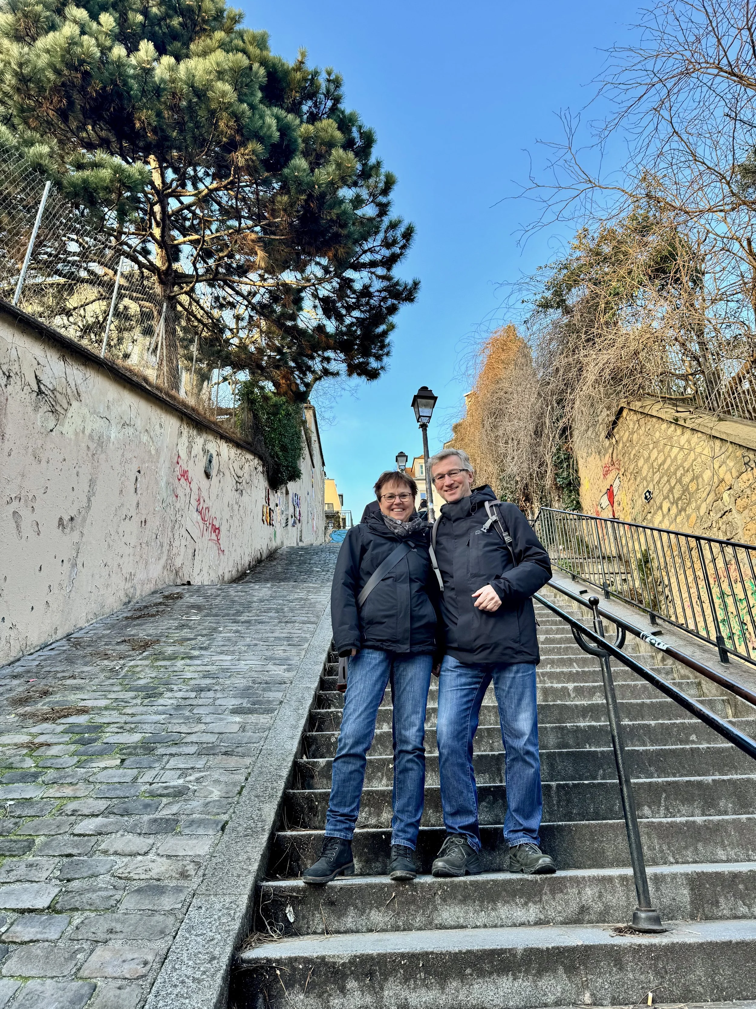 A smiling couple standing on outdoor stairs in a city, dressed in jackets and jeans, with trees and street lamps in the background.