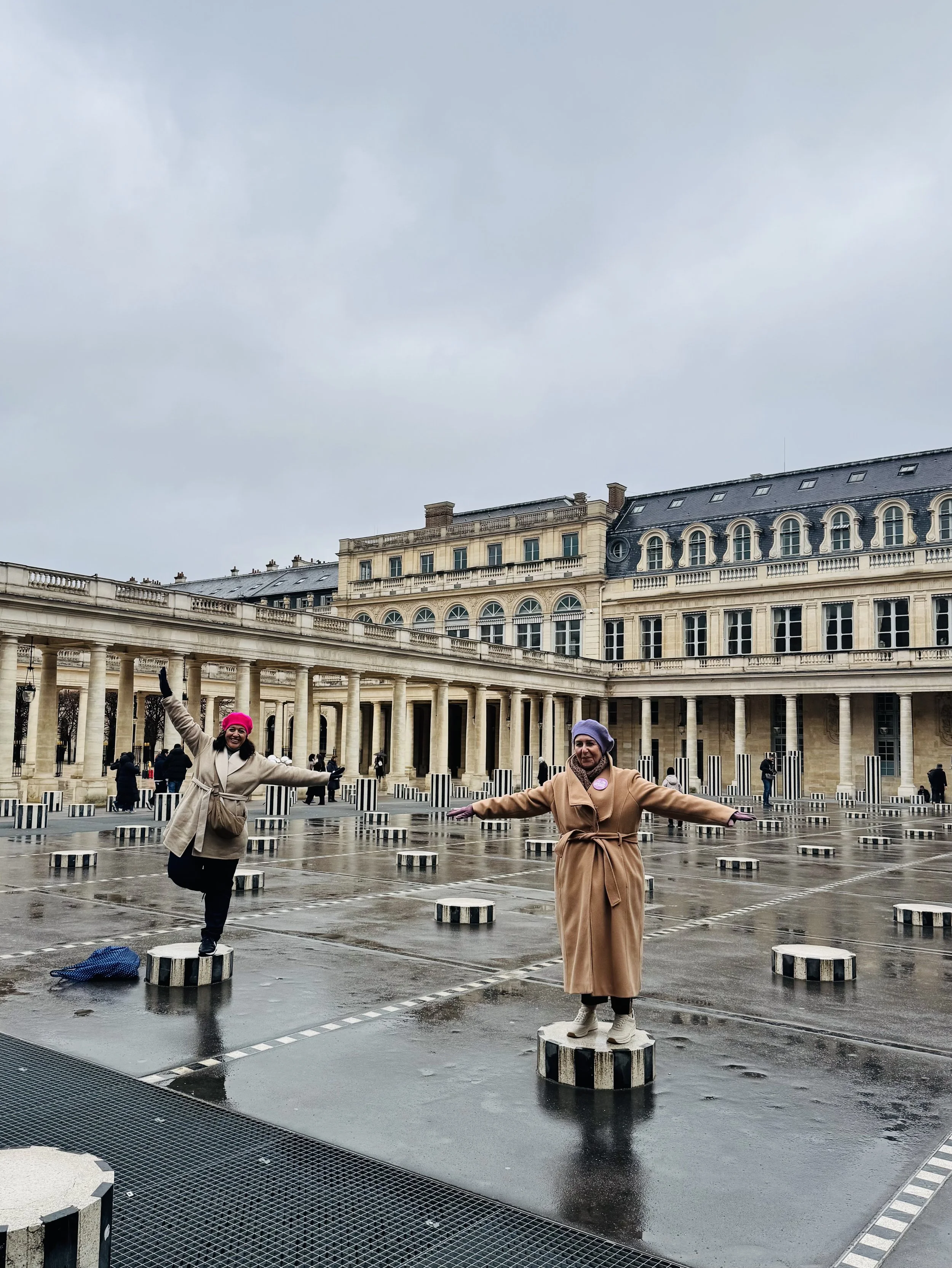 Two women standing on striped black and white circular bollards in an outdoor plaza with historic building in the background, on a rainy day.