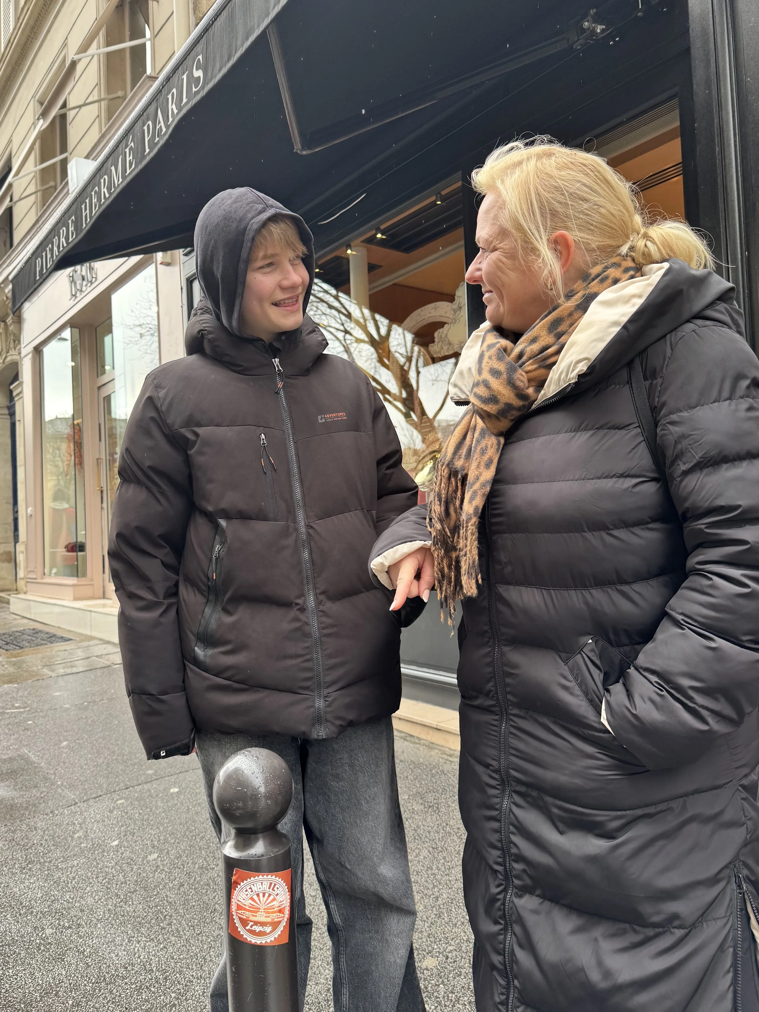 A young man and an older woman are standing outside on a city street, engaging in a warm hug and conversation. They are both bundled in winter jackets, with the woman wearing a leopard print scarf. The background shows storefronts and a curb with a b