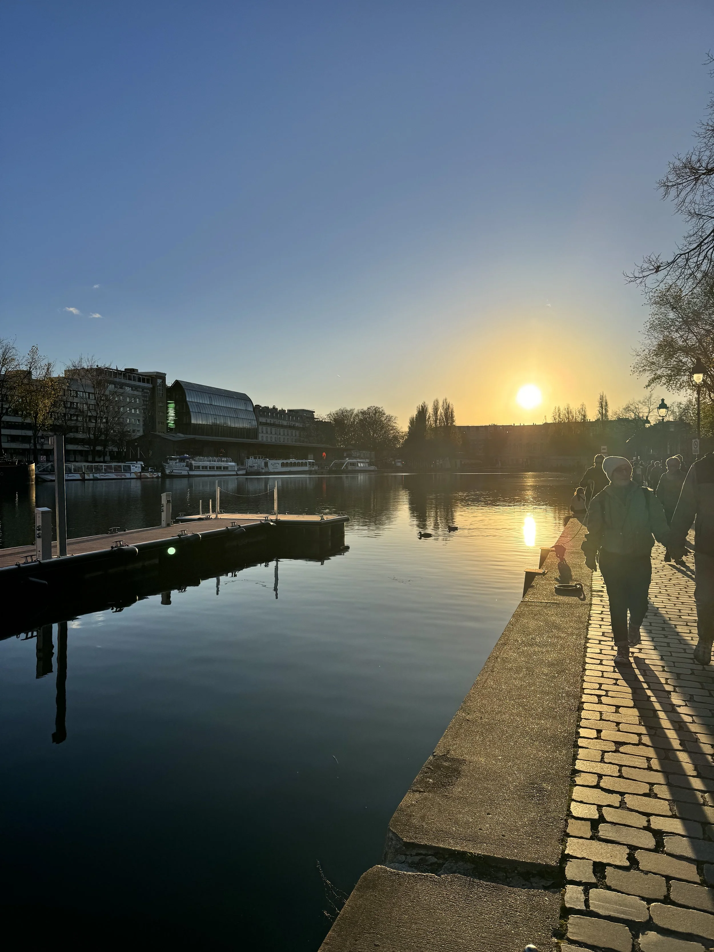 People walking along a riverside promenade during sunset, with boats docked by the water and a modern building in the background.