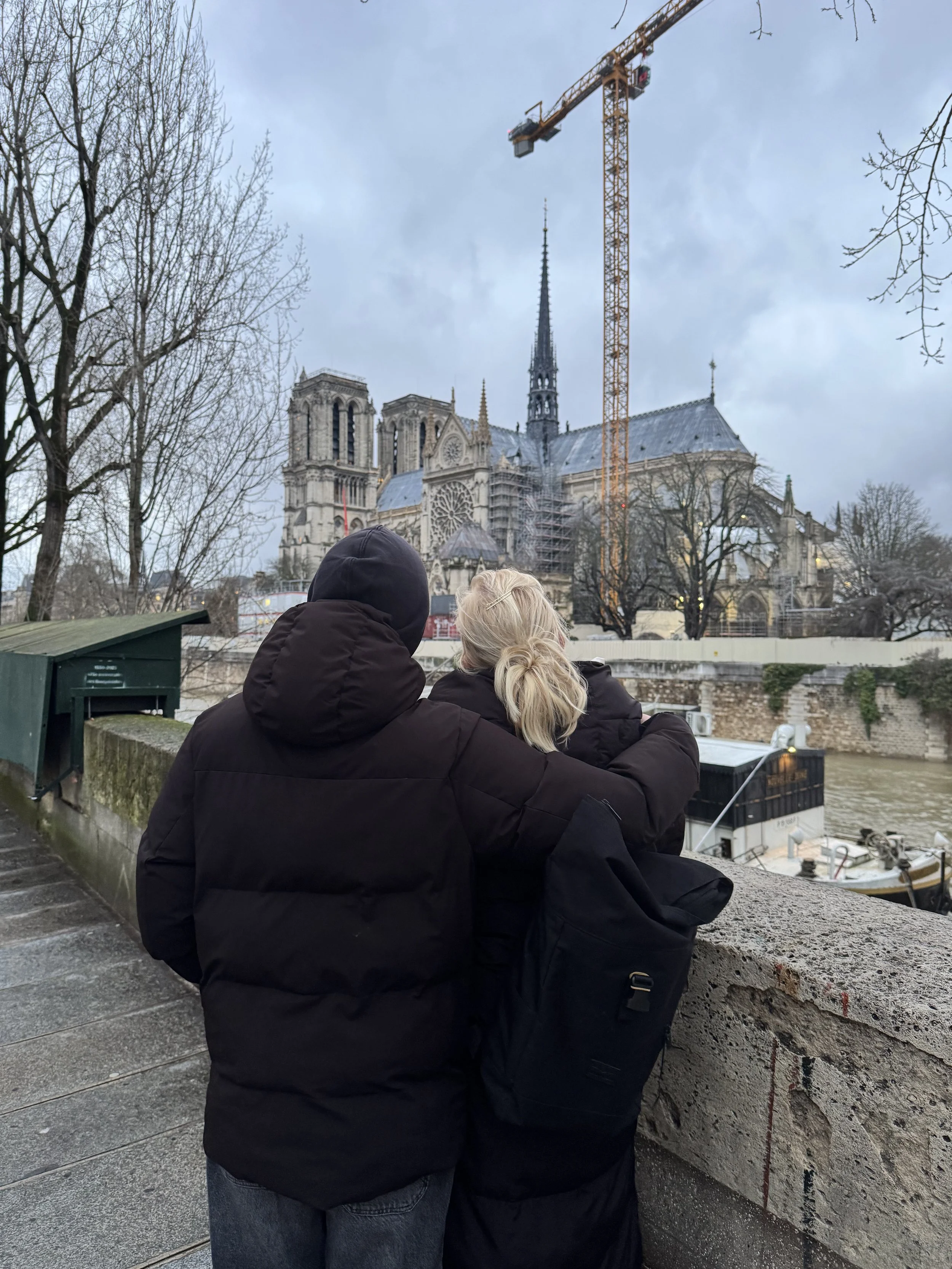 Two people in dark jackets embrace on a bridge overlooking the Seine River with Notre-Dame Cathedral in the background in Paris.