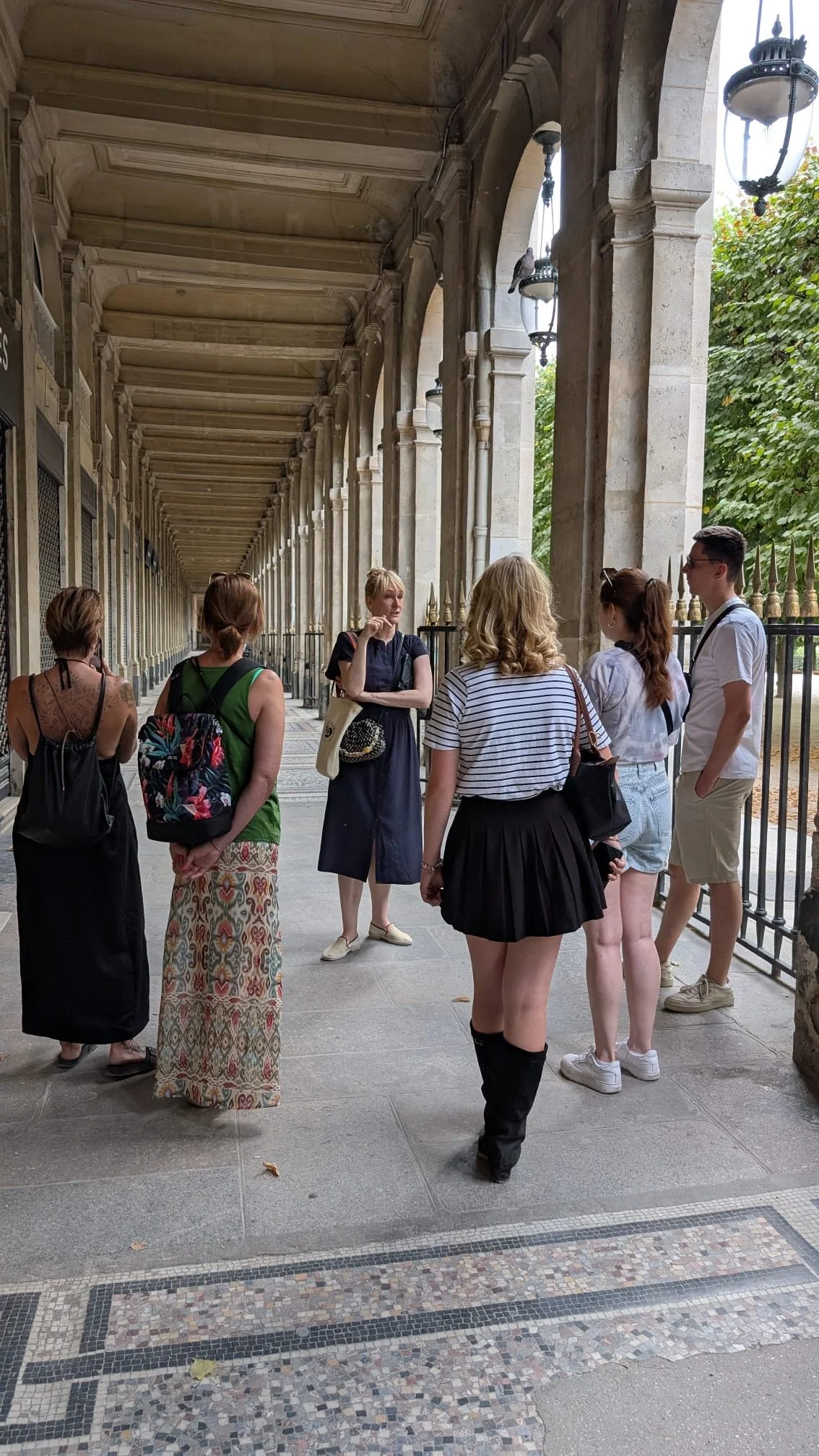 Gruppe von Menschen, hauptsächlich Frauen, in einem überdachten, historischen Arkadengang, die miteinander sprechen, einige tragen Rucksäcke, es ist sonnig, Bäume sind sichtbar.