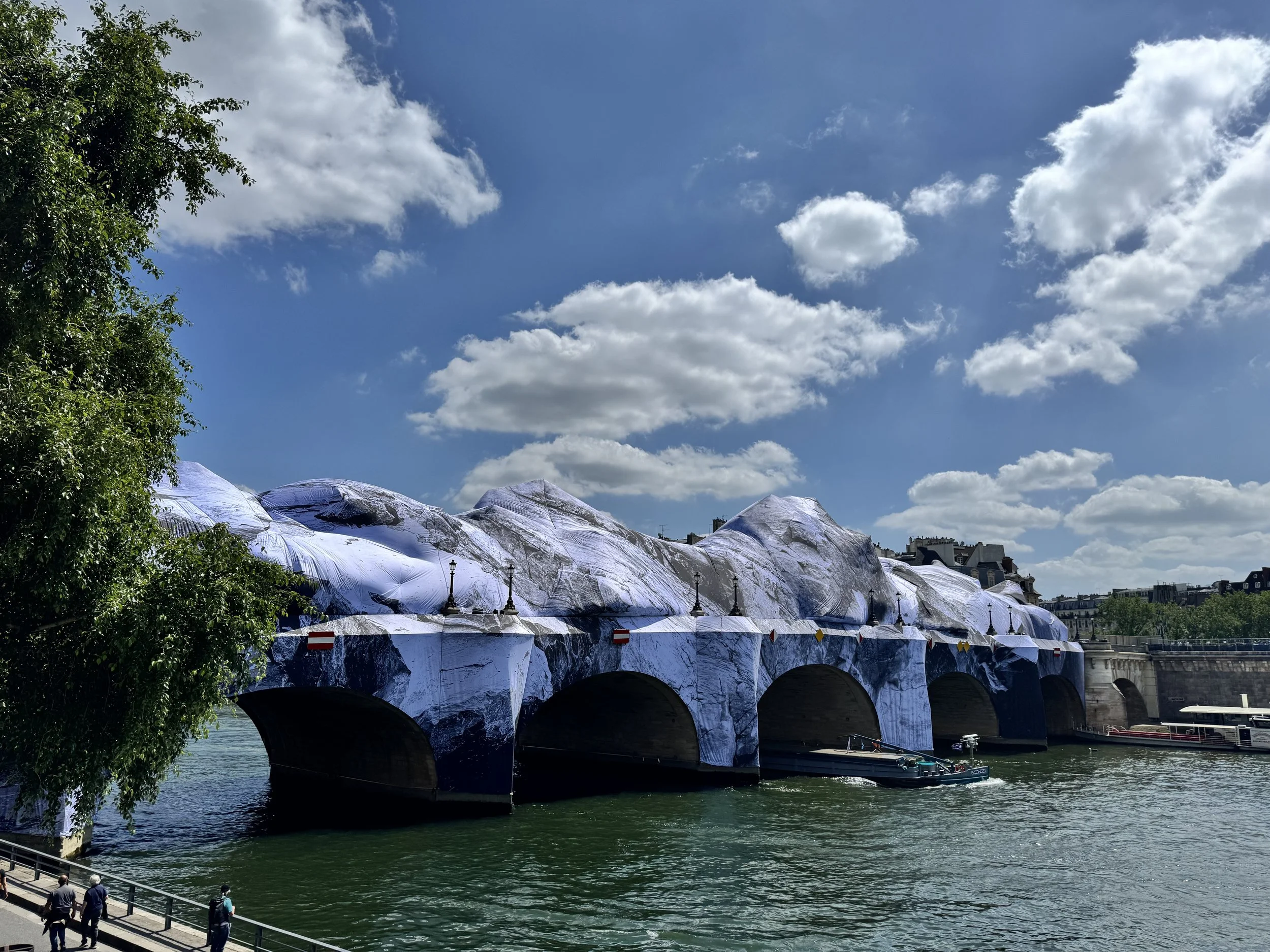 Uberspannte Strukturen mit silberner Abdeckung entlang eines Flussufers, eine Gruppe von Menschen spaziert auf dem Gehweg, im Hintergrund Gebäude und grüne Bäume, blauer Himmel mit Wolken.