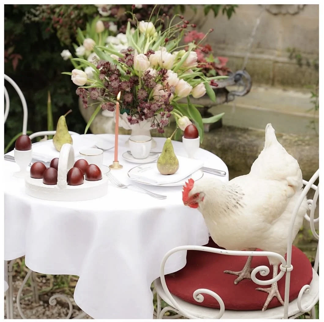 Ein gedeckter Gartentisch mit Blumenstrauß, Eiern, Pears, Kerze und Huhn auf einem Stuhl