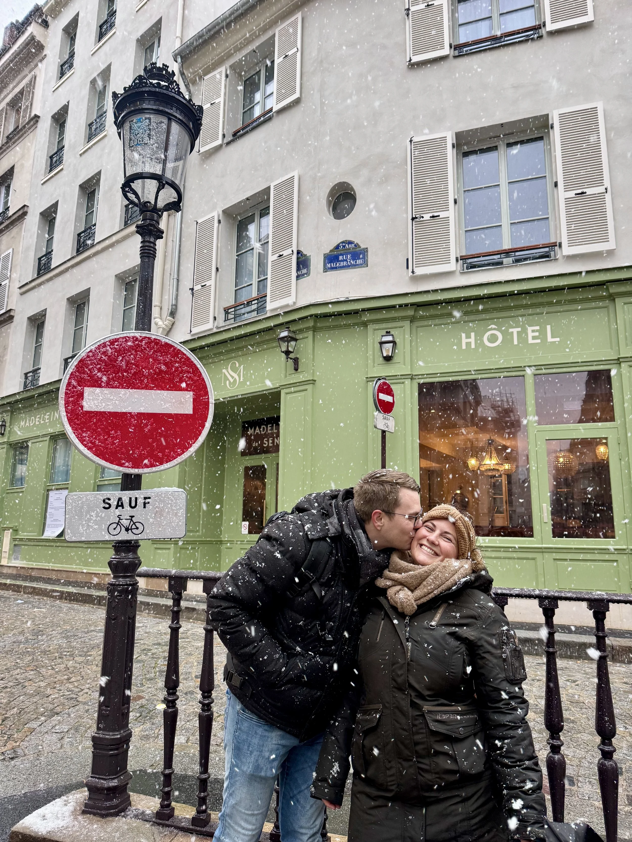 A man kisses a woman on the cheek in front of a green hotel with snow falling, on a European city street with signs including a red no entry sign and a bicycle exception sign.