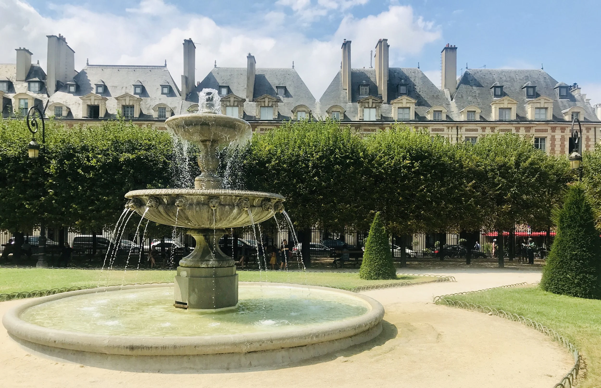 Eine Springbrunnen in einem Park mit grünen Büschen, im Hintergrund alte Gebäude mit grauen Dächern und Schornsteinen