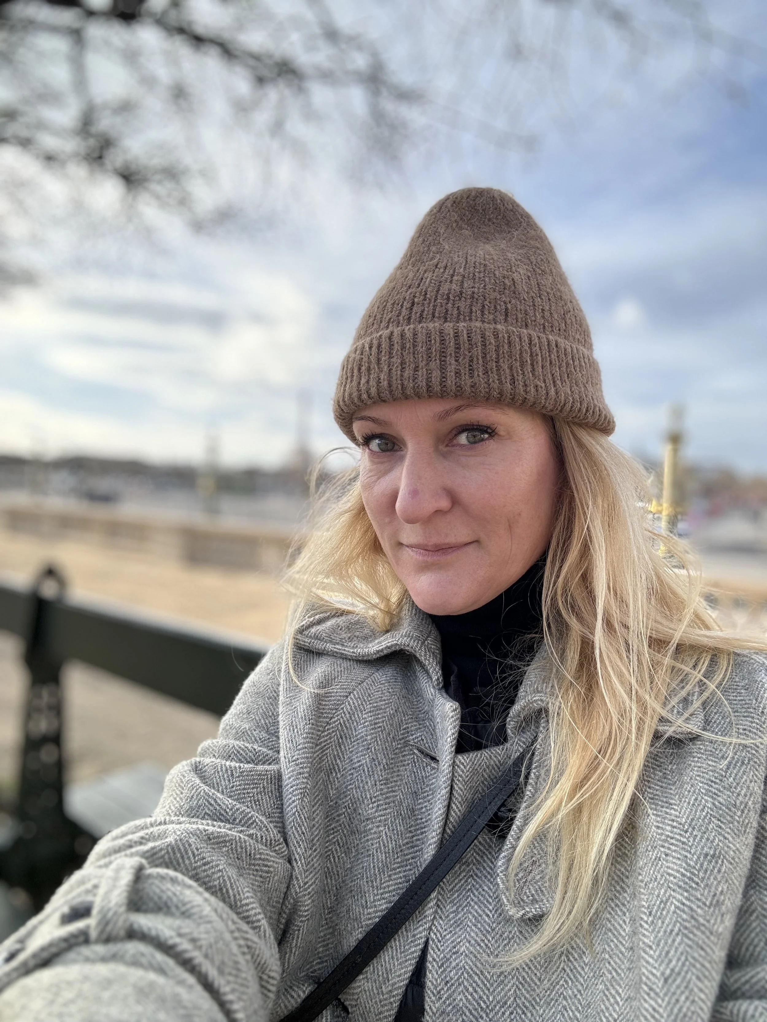 A woman with long blonde hair, wearing a brown knit beanie and a gray checkered coat, sitting on a bench outdoors with a blue sky and trees in the background.