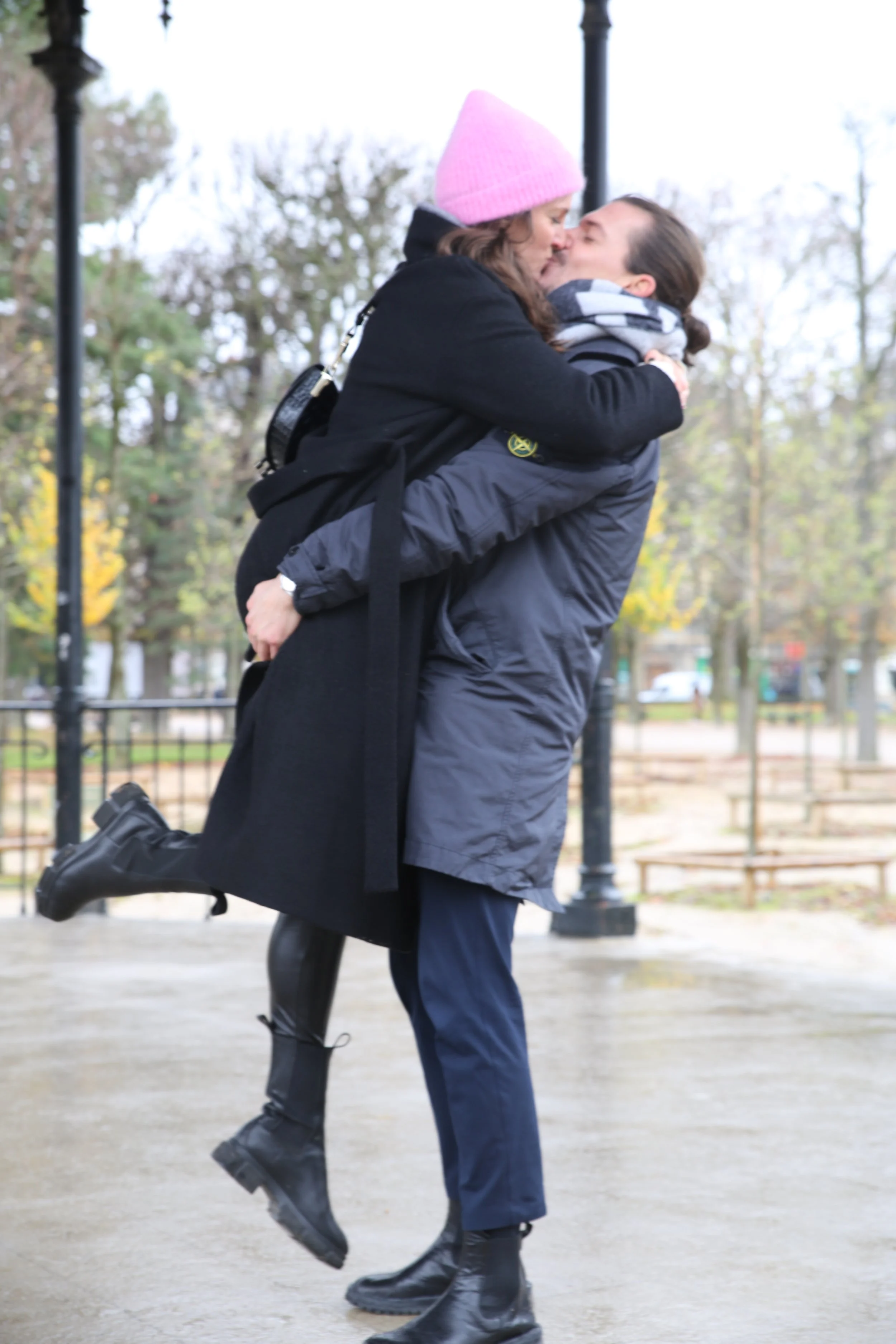 Man lifting woman in an outdoor park, both dressed in warm coats and boots, sharing a kiss.