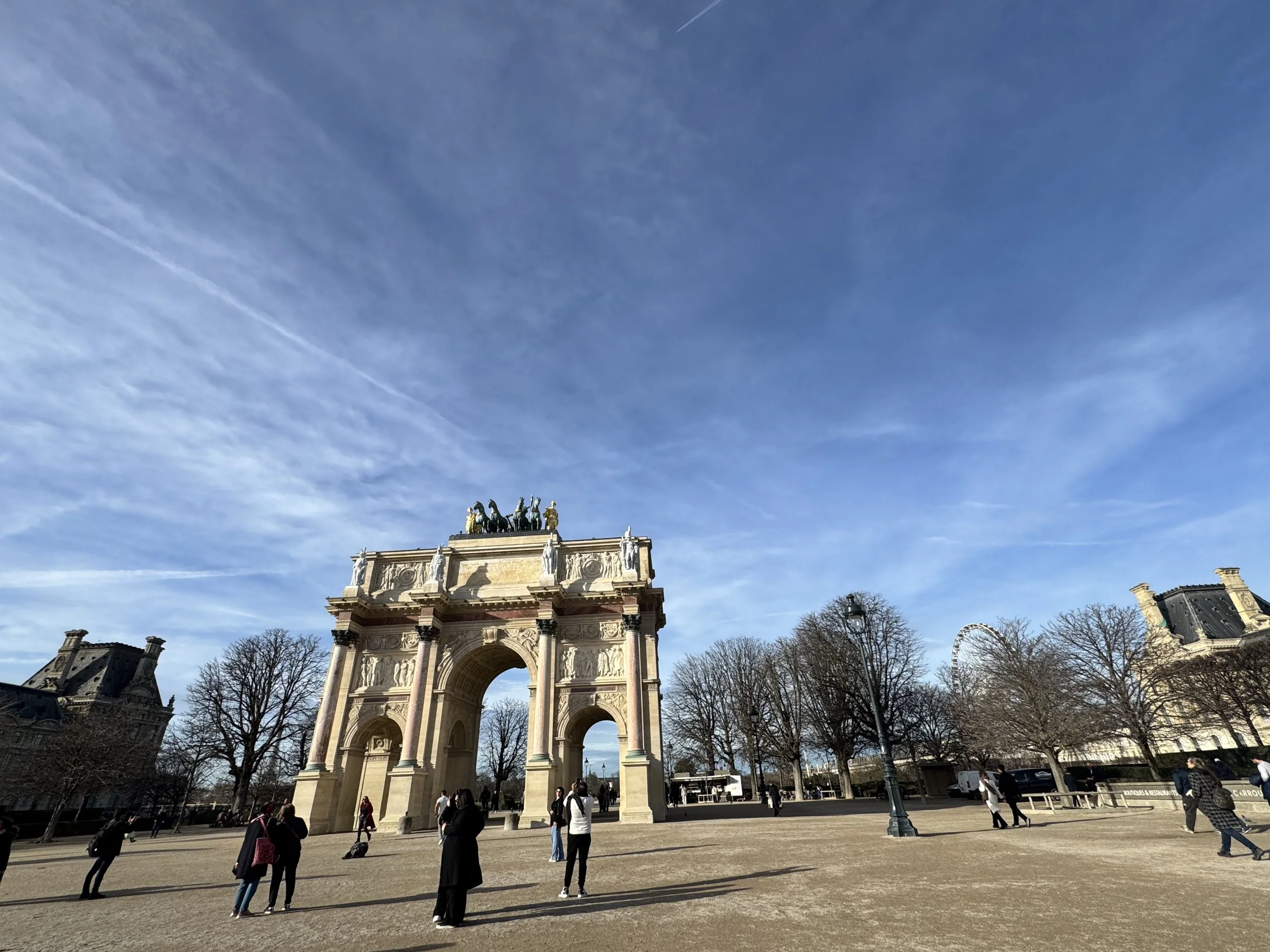 People walking near the Arc de Triomphe in a park on a clear, blue sky day.