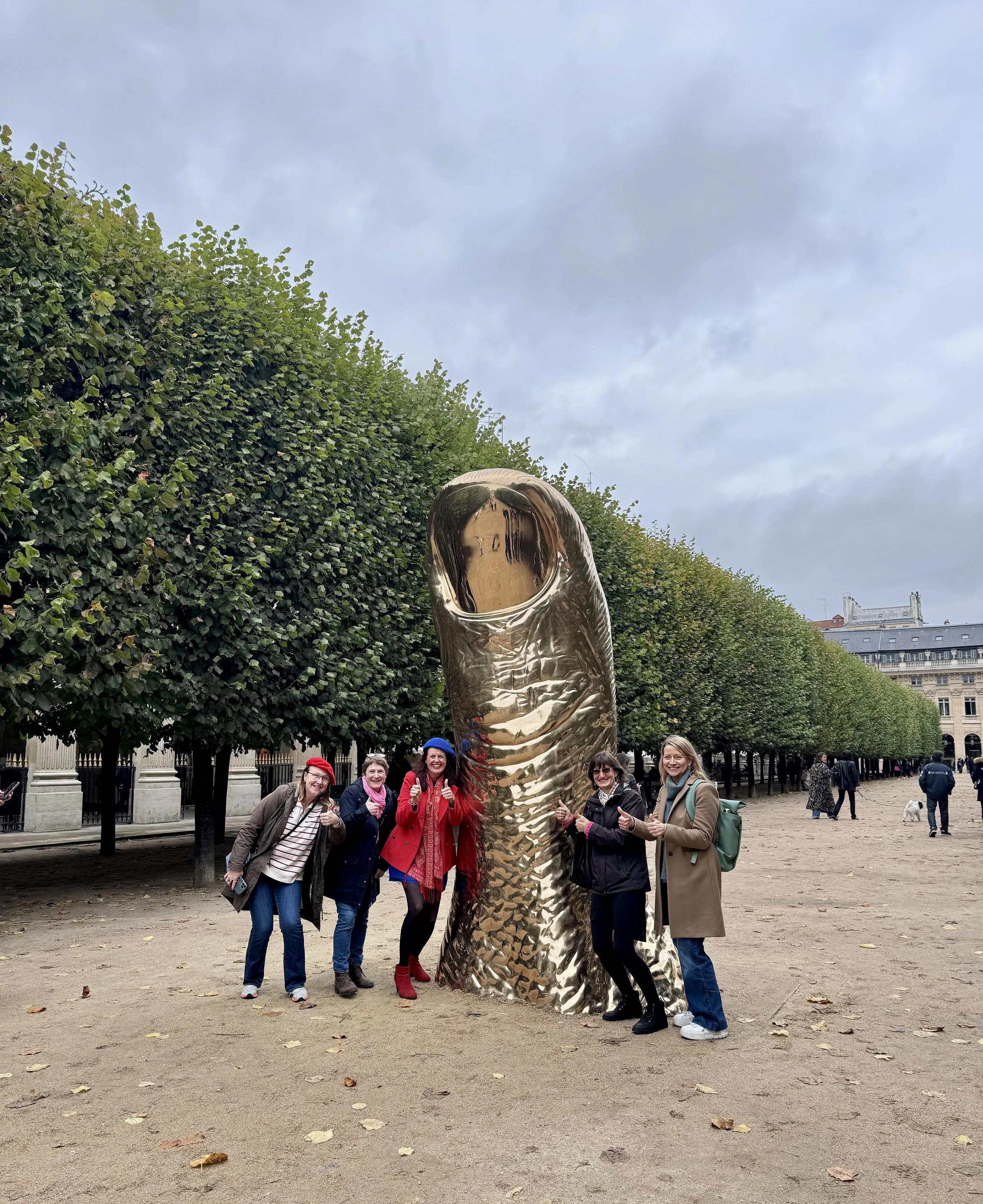 A group of five women standing around a large, shiny, gold sculpture of a thumb, in an outdoor park with trees and a cloudy sky.