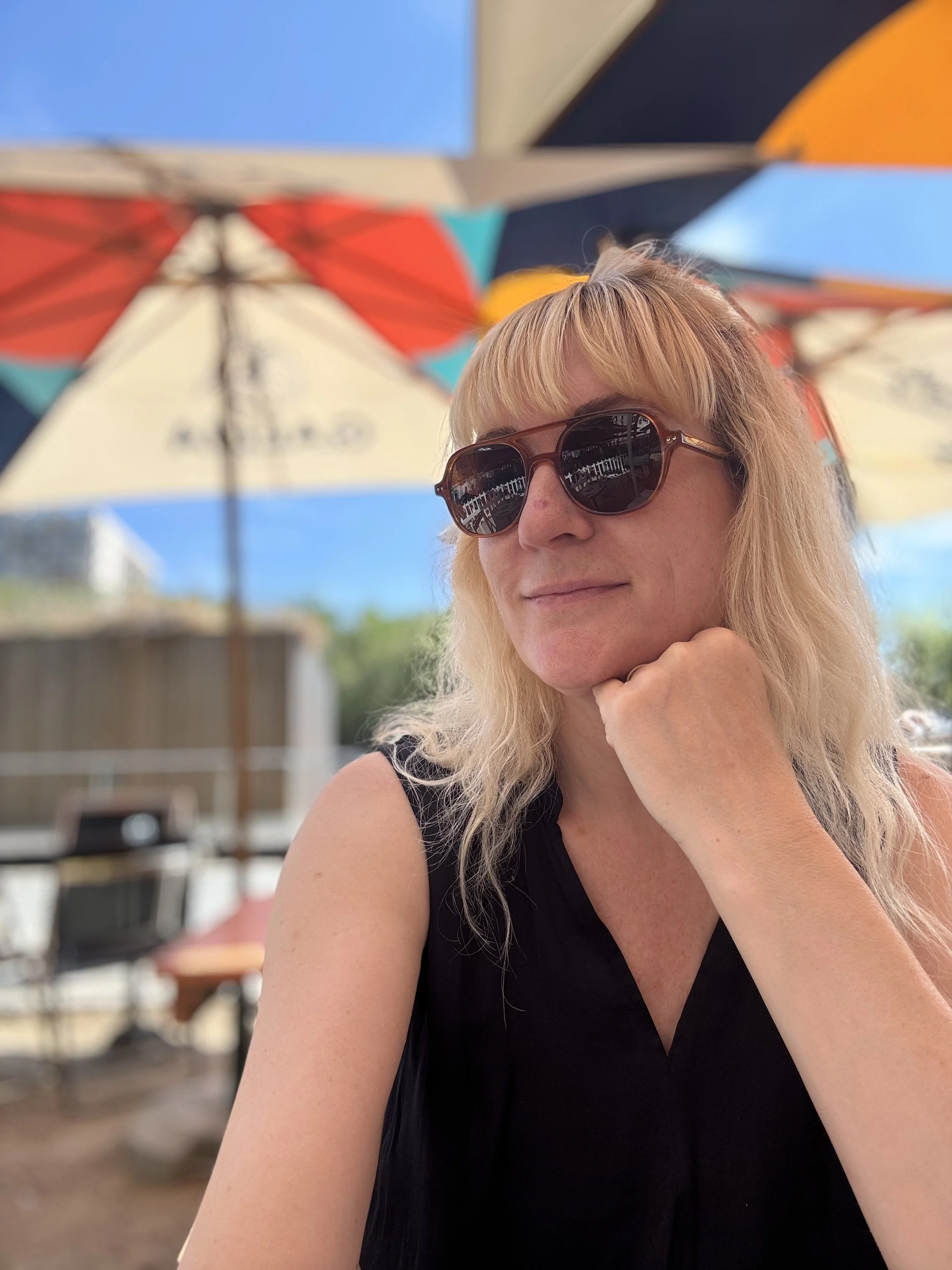 Eine Frau trägt Sonnenbrille und sitzt unter einem bunten Sonnenschirm bei blauem Himmel draußen.