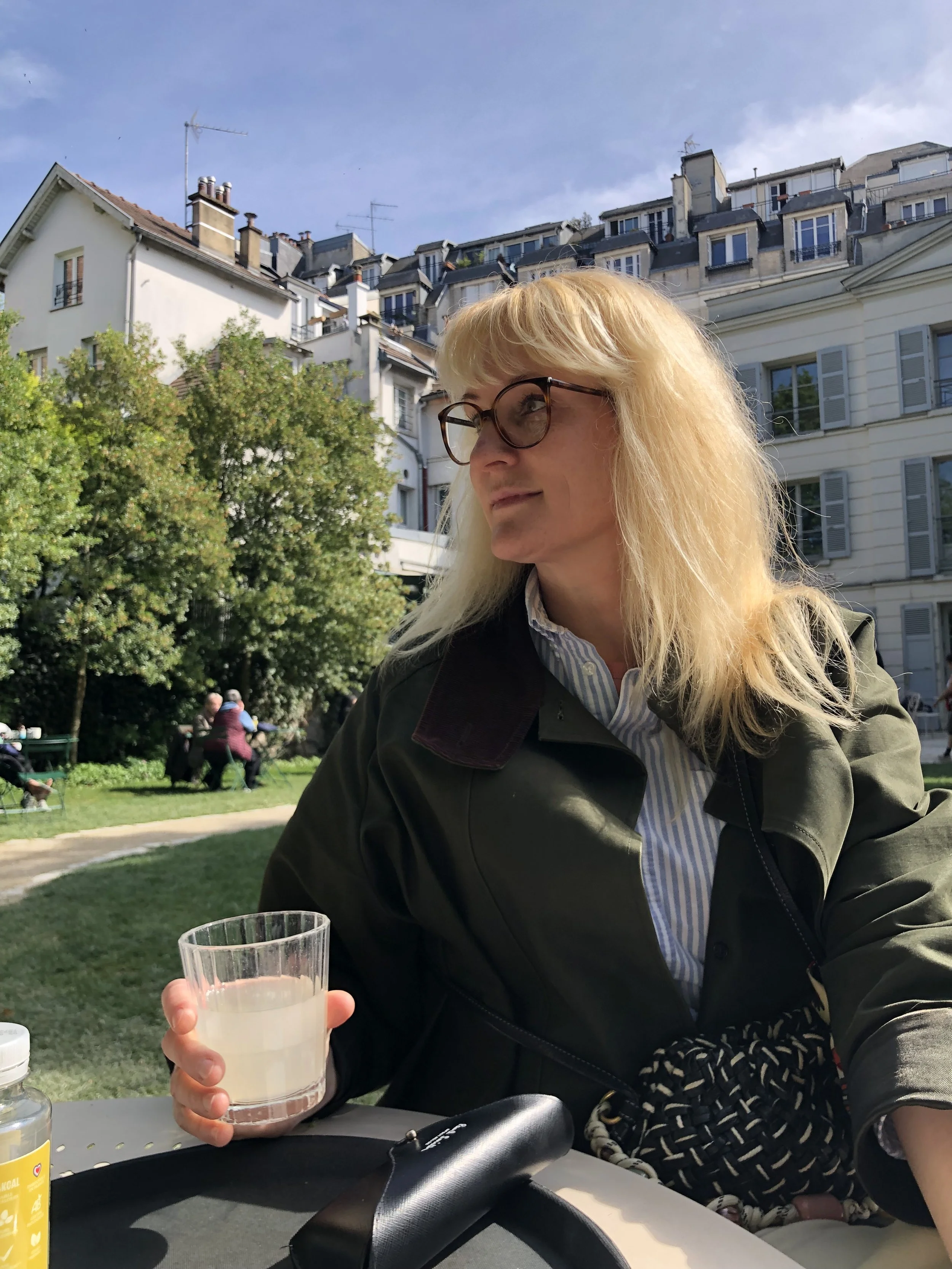 Eine Frau mit blonden Haaren, Brille und dunkler Jacke sitzt im Freien in einem Park oder Garten, hält ein Glas mit einem weißen Getränk, im Hintergrund sind Bäume, Häuser und einige Menschen zu sehen.