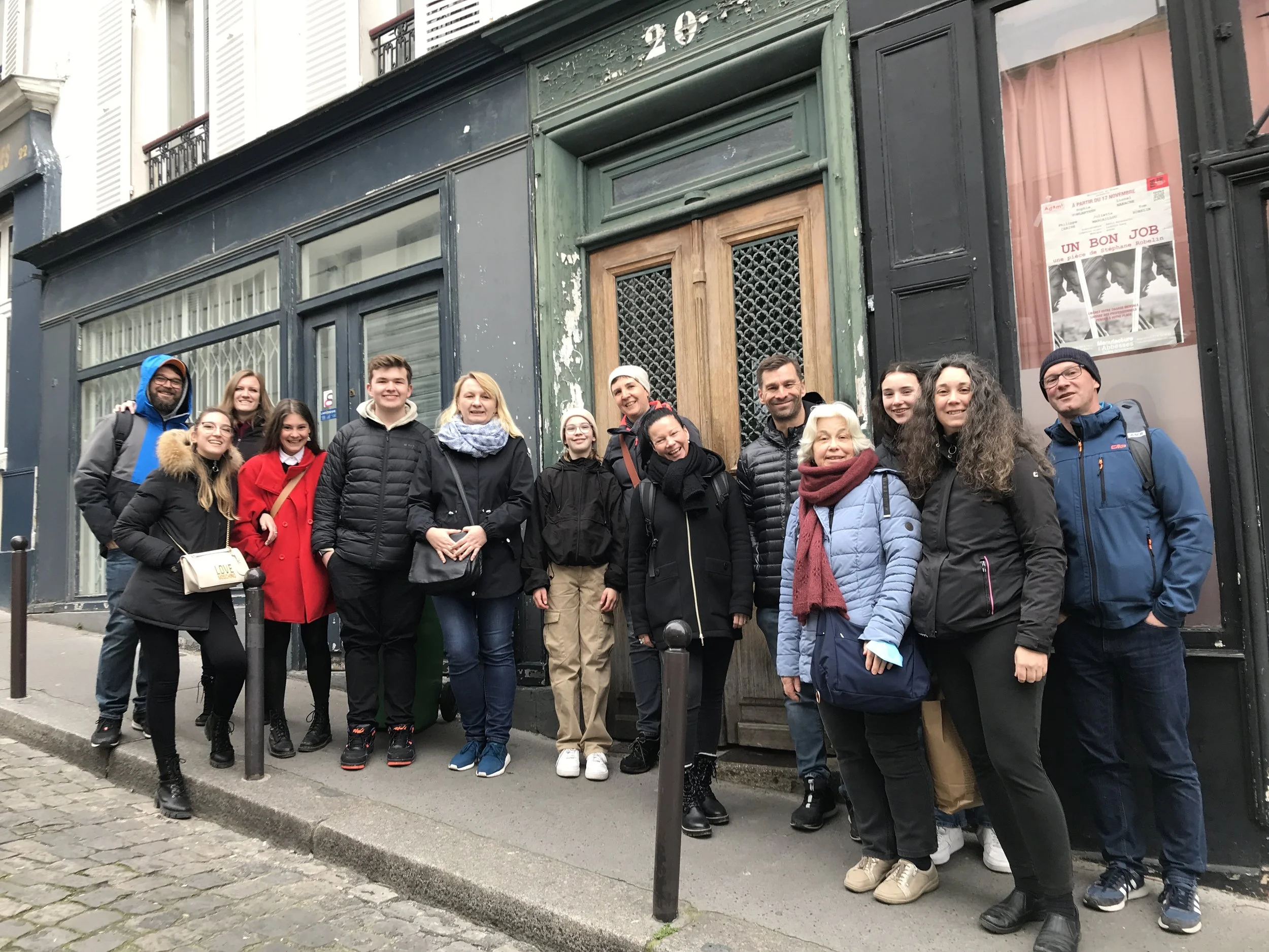 Group of 14 people standing on a sidewalk in front of a dark green and black building, posing for a photo. The group appears to be tourists or friends, dressed in casual winter clothing.